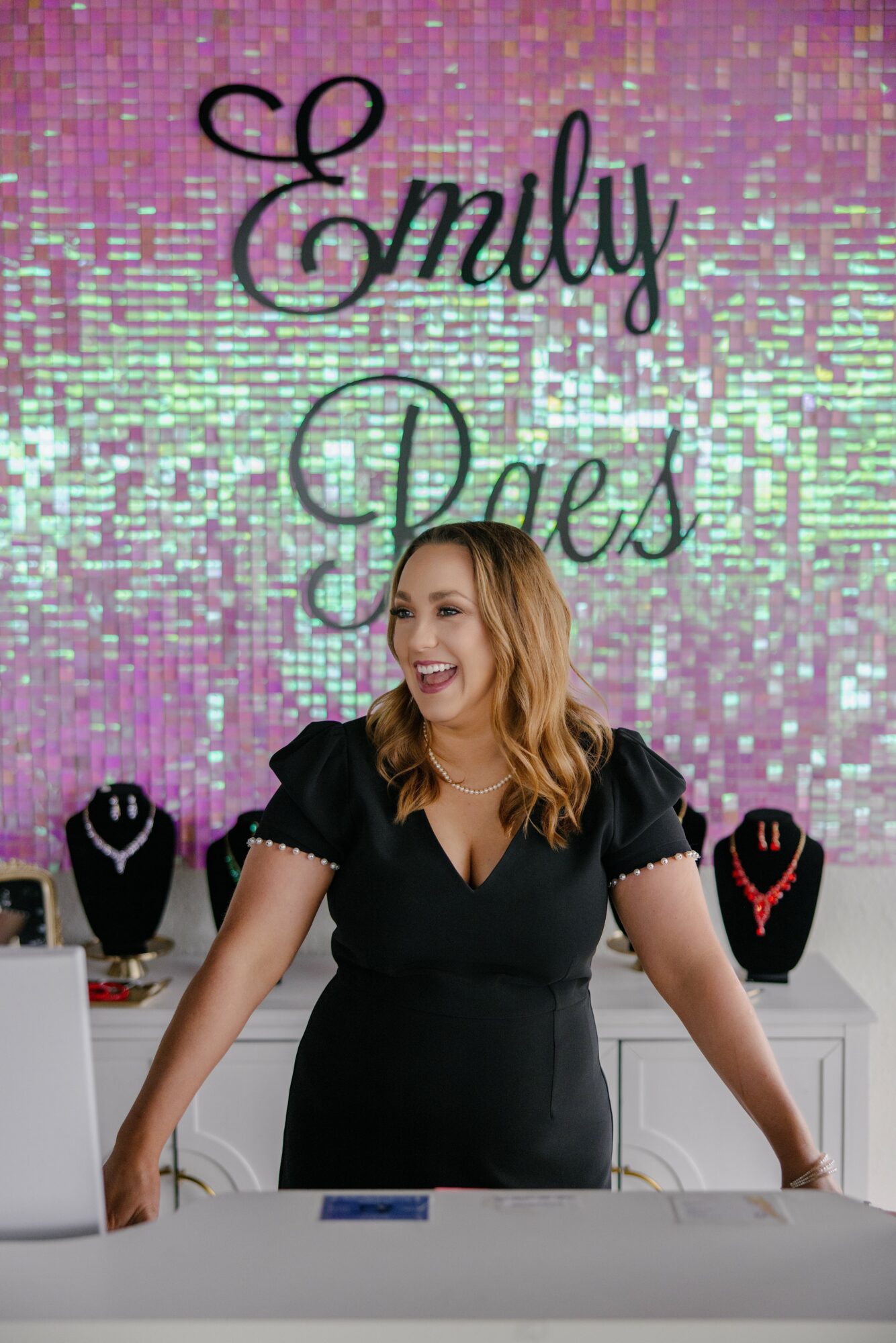 Woman smiling at a celebration with a colorful, glittery backdrop reading 'Emily' and 'Packs'.