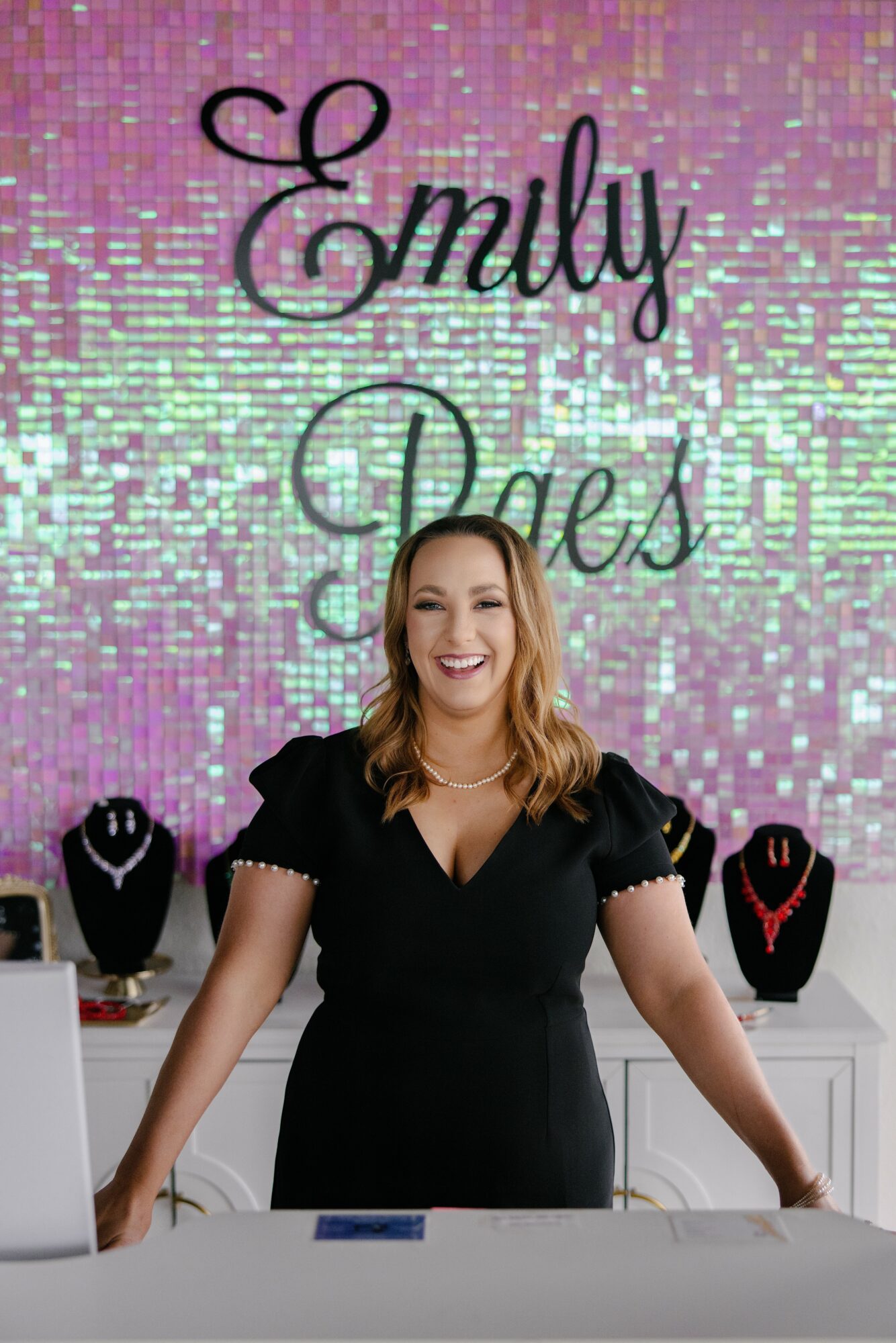 Woman smiling at jewelry display with a colorful, sparkly background and the words 'Emily' and 'Dax' above.