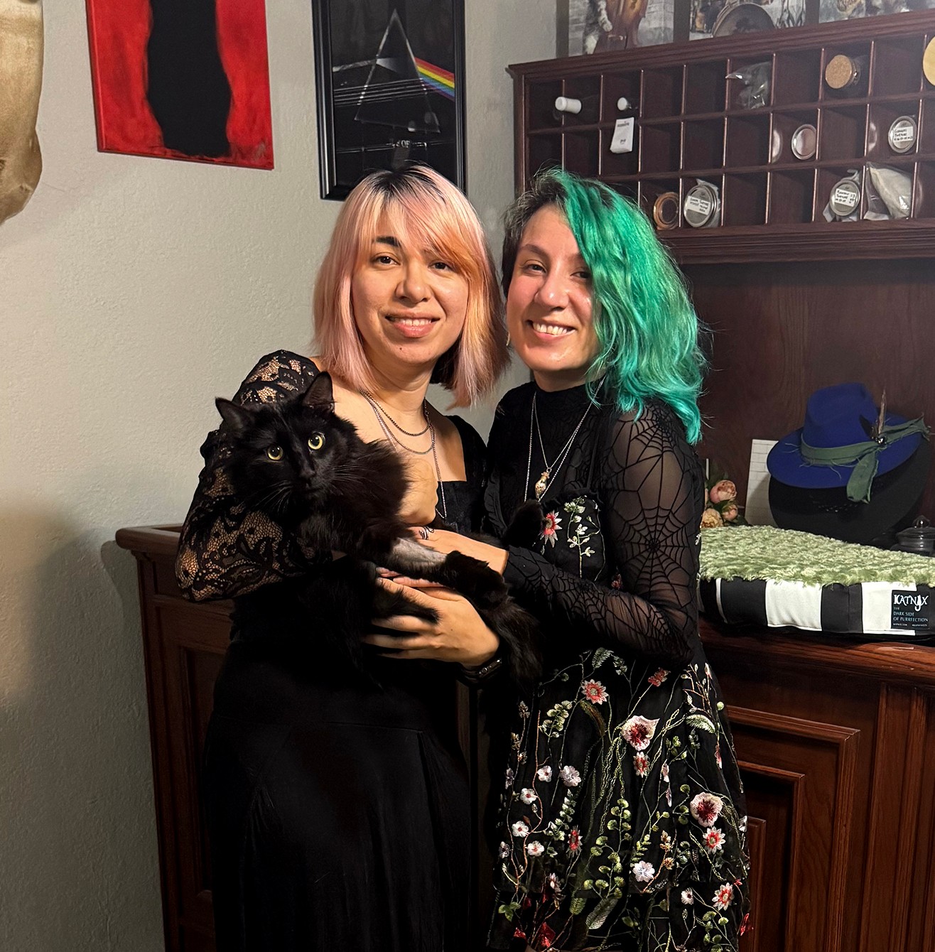 Two smiling women with colorful hair holding a small dog indoors, with shelves and artwork in the background.