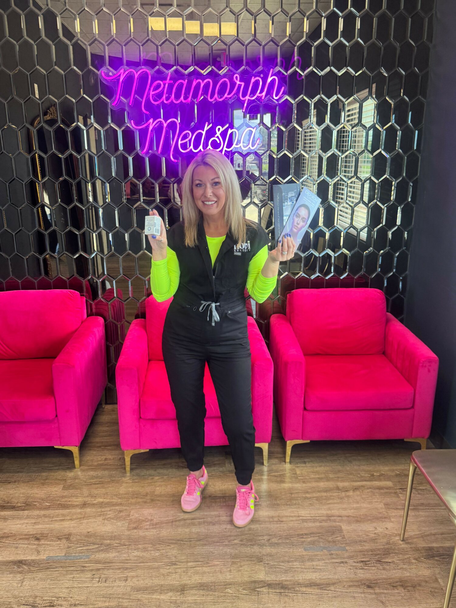 Person with blonde hair sitting on a pink chair, holding a phone, in front of a neon sign and black honeycomb wall.
