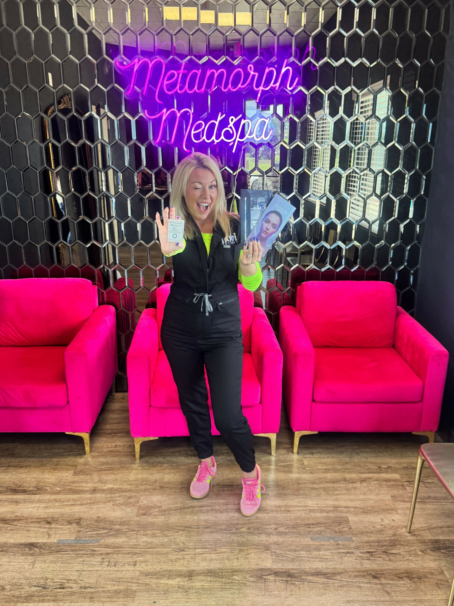 Woman taking a selfie in a room with pink furniture and a neon sign, standing in front of a pink sofa and chair.