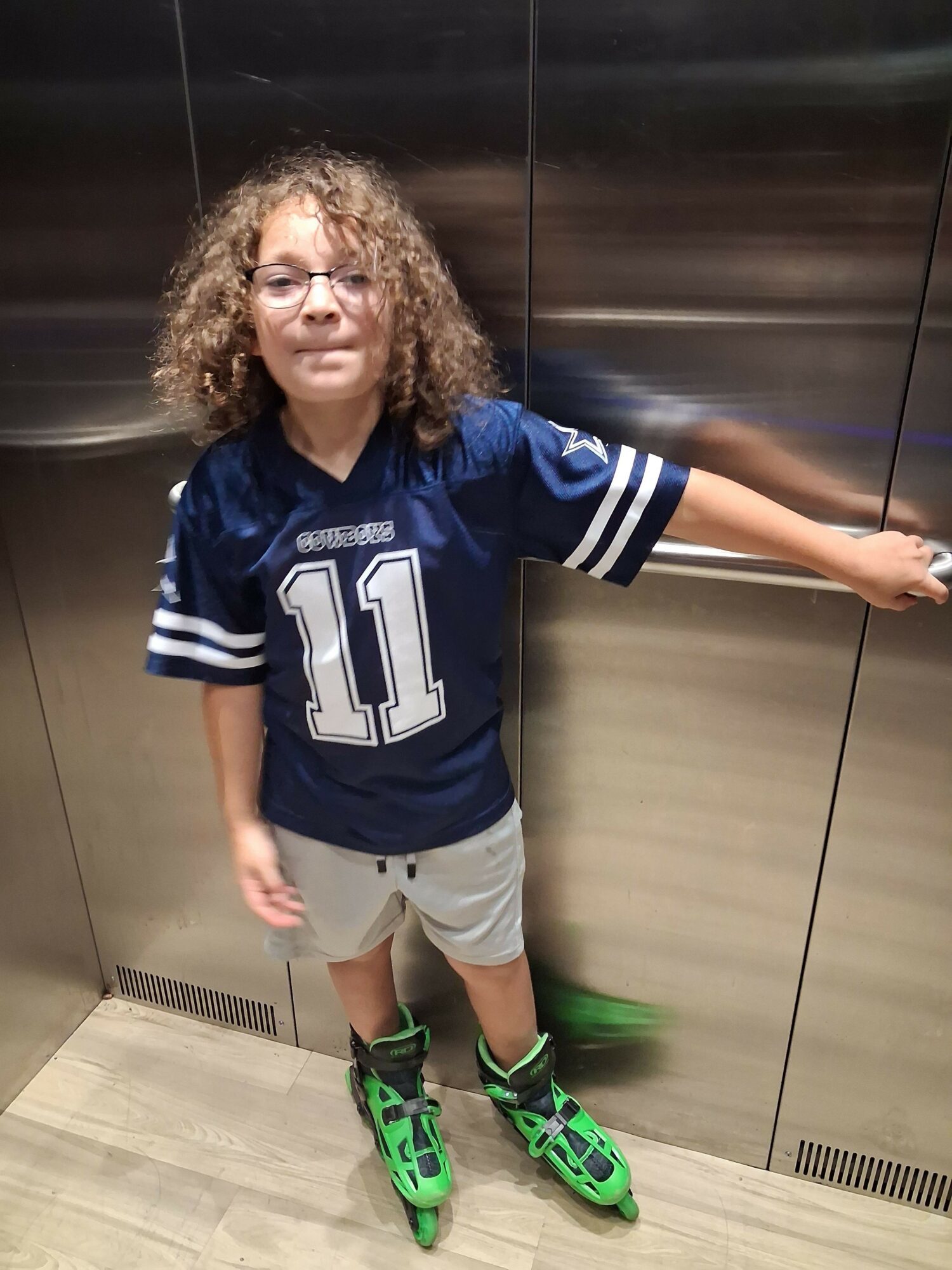 Young girl with curly hair smiling, wearing a blue sports jersey with number 11, gray shorts, and green shoes, standing indoors.