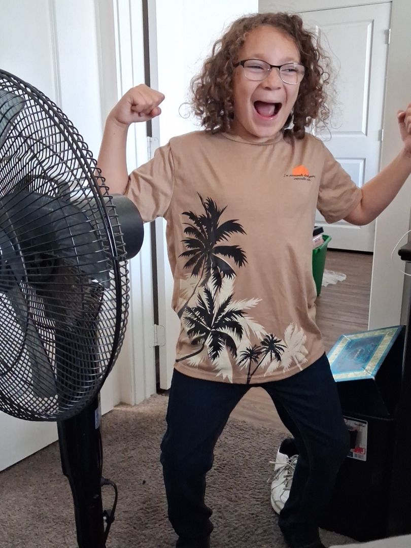 Person with curly hair and glasses celebrating indoors, wearing a beige shirt with palm tree print, raising a fist.