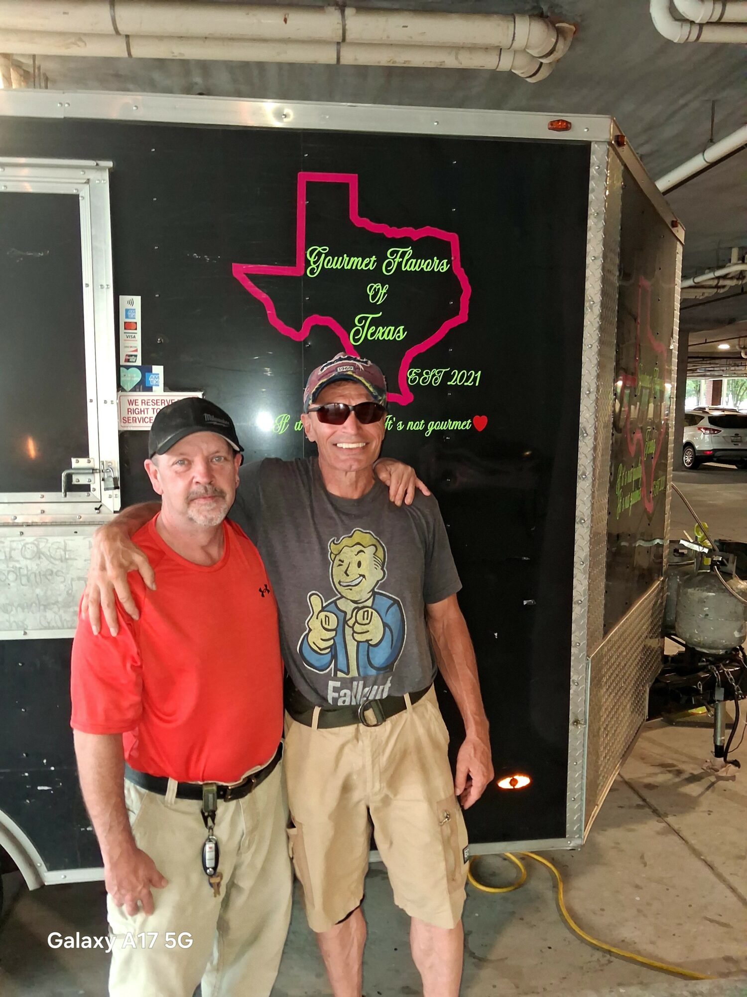 Two men standing together, one with arm around the other, in front of a black trailer with Texas-shaped sign and text, indoors.