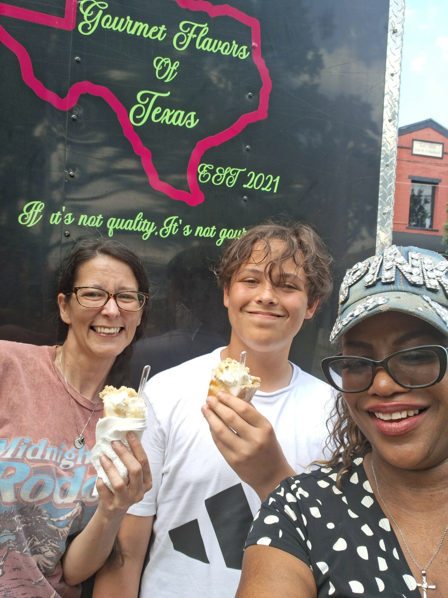 Three women smiling and holding ice cream cones, standing outdoors near a blackboard with green and pink writing.