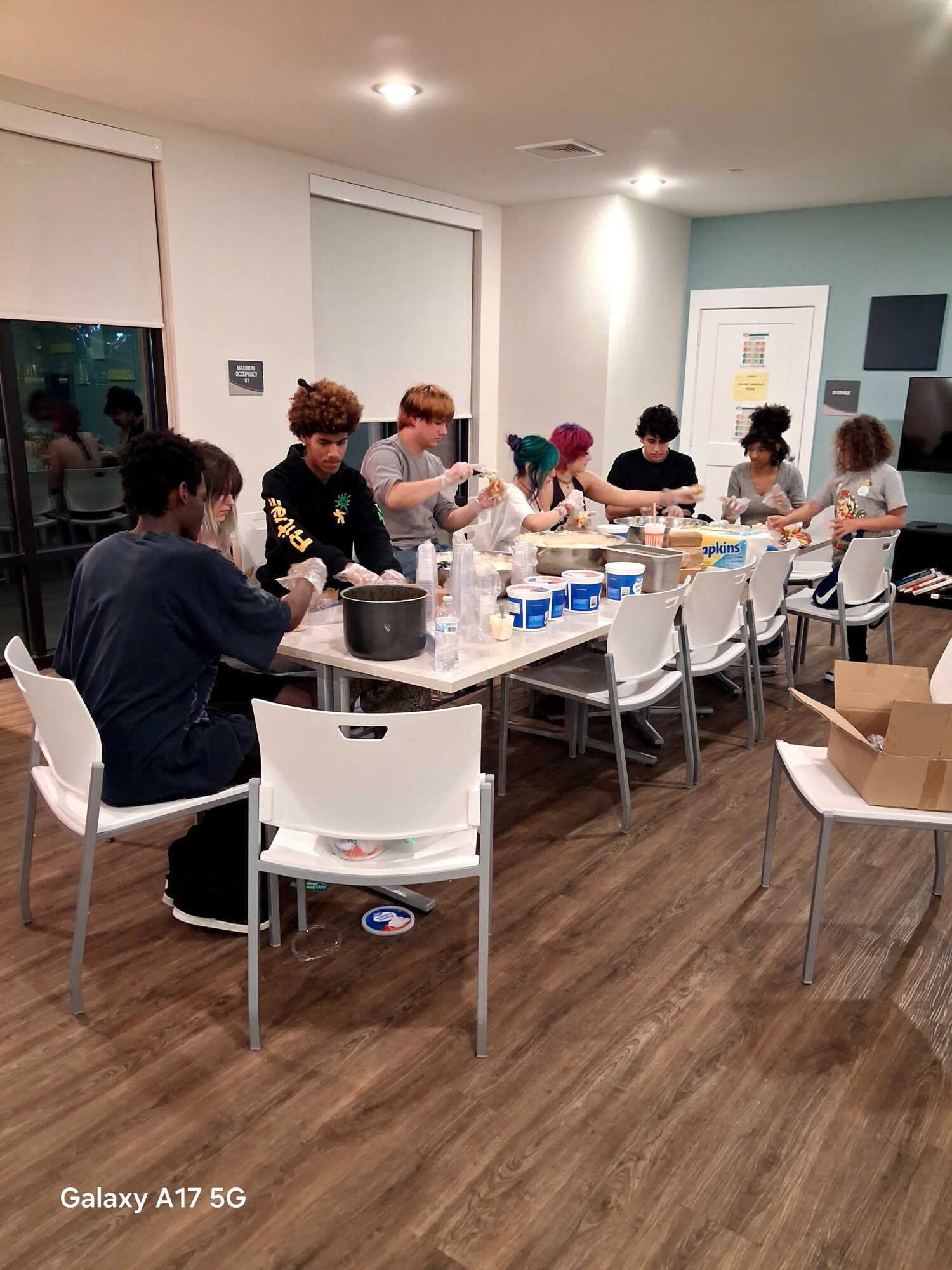Group of people sitting at a long table with cups and containers, in a room with white and blue walls, wooden floor.