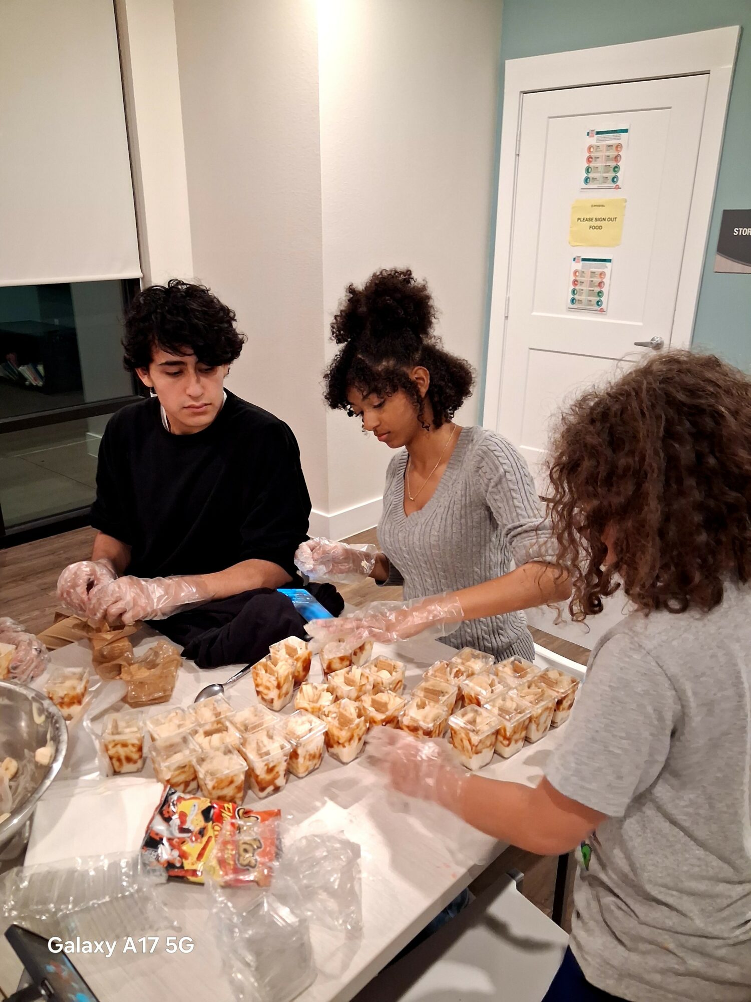 Three children with curly hair working together at a table, packaging baked goods in a kitchen. There are snacks and baked items on the table.