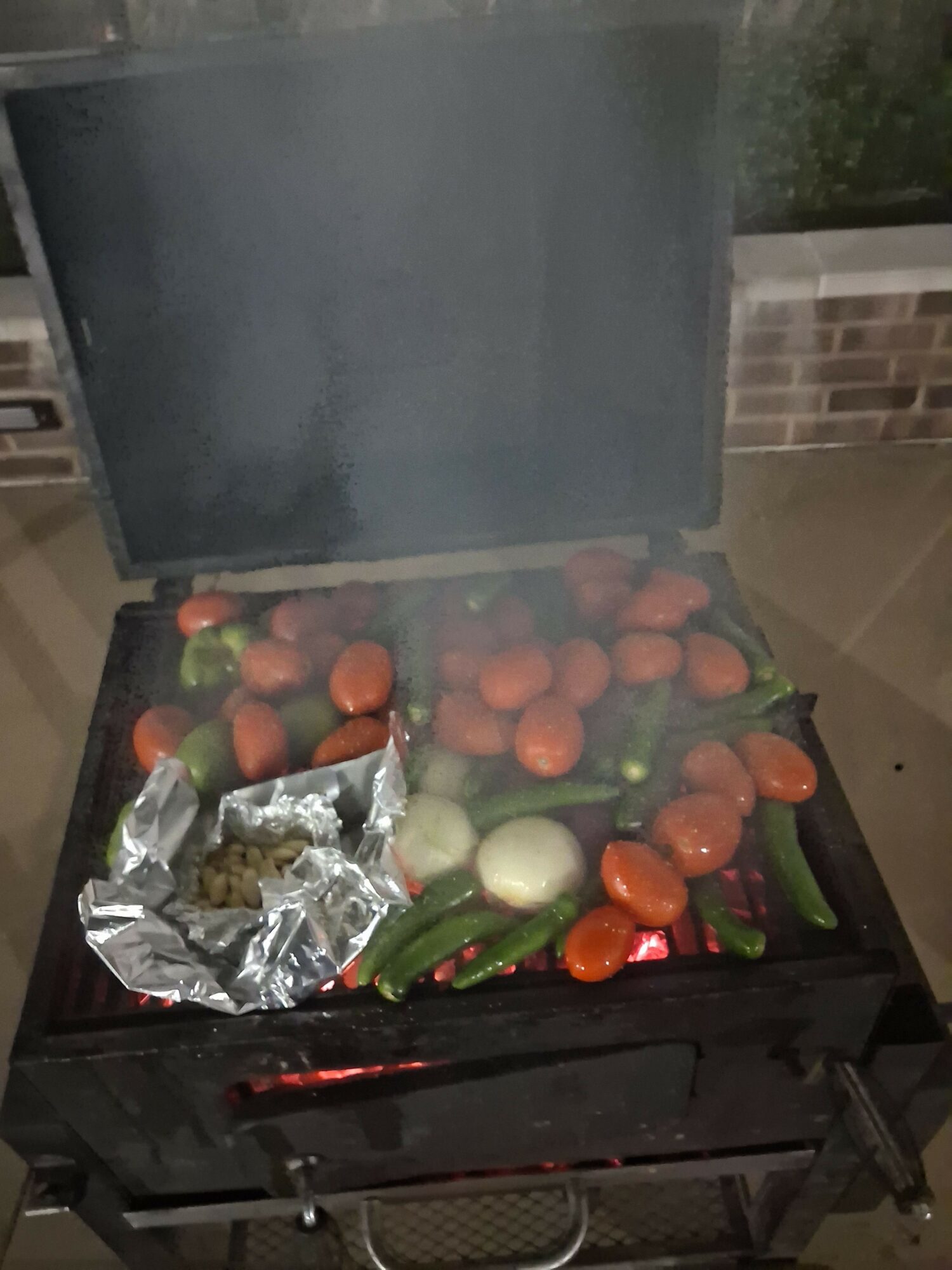 Vegetables including tomatoes, cucumbers, onions, and peppers on a grill with foil-wrapped food, viewed from the side.