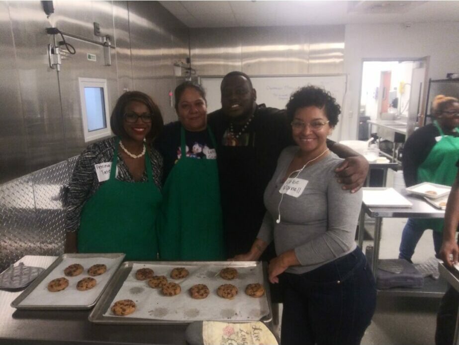 Three people standing behind a table with baked goods in a kitchen, smiling at the camera.