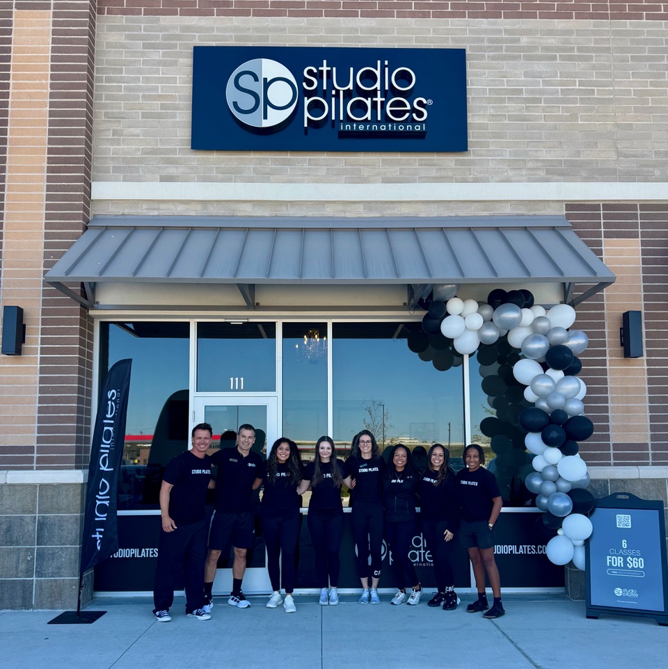 Group of seven people standing outside Studio Pilates, with balloons and signage, in front of a glass door and window.