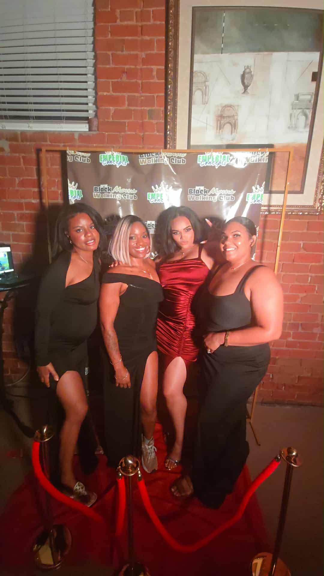 Four women in black and red dresses posing together indoors with a brick wall and window behind them.