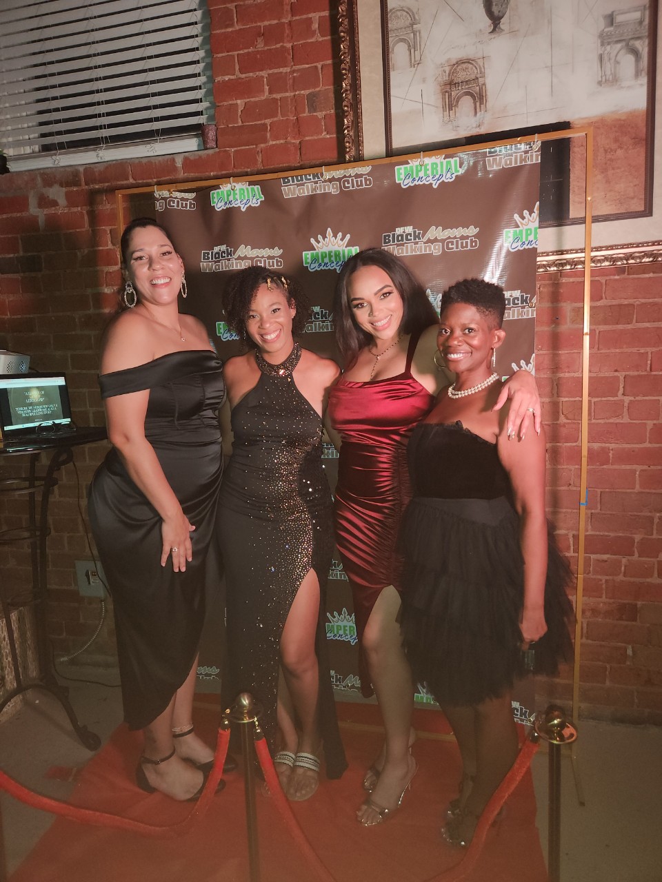 Four women in elegant dresses pose together in front of a backdrop with logos, smiling at the camera.