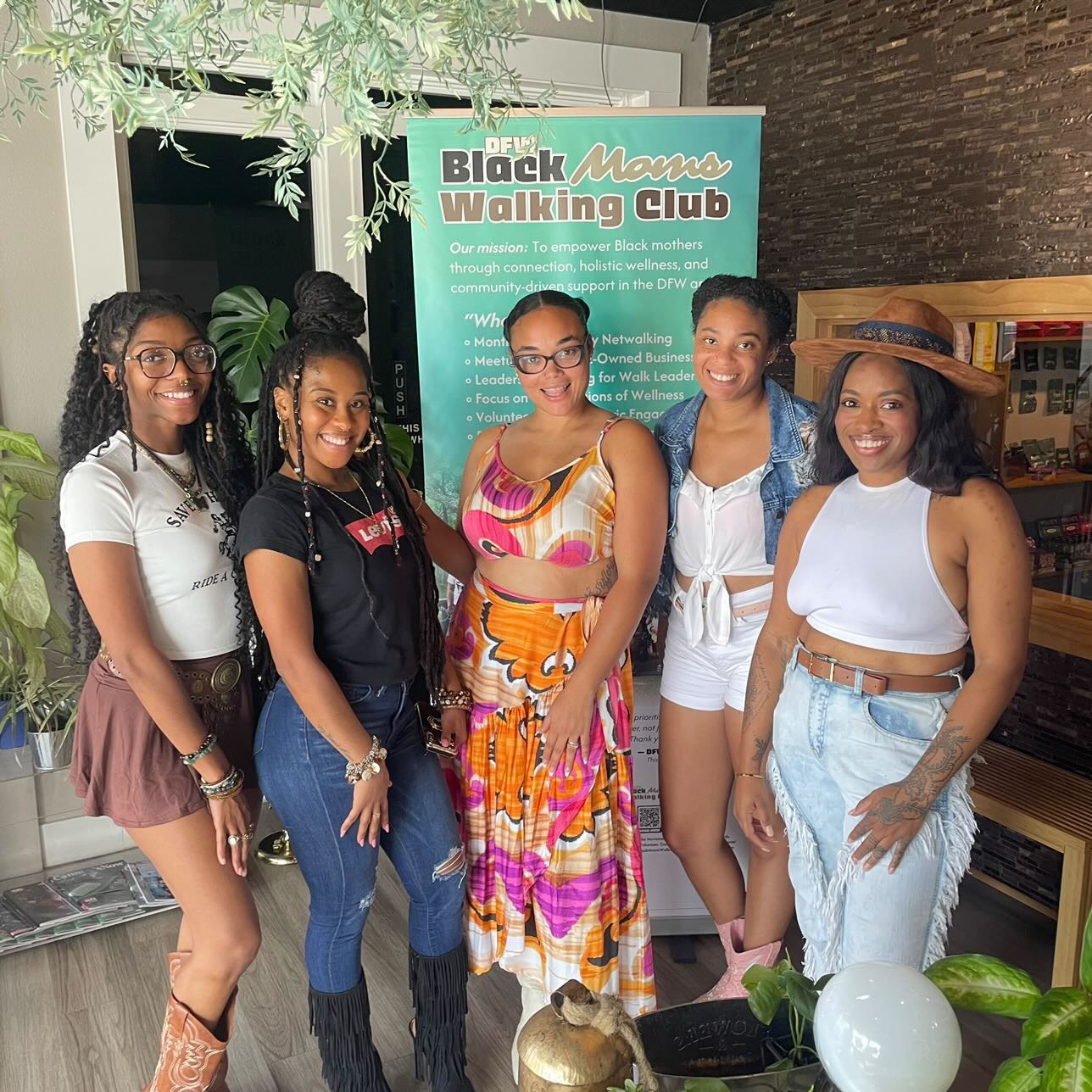 Six women standing together indoors, smiling, with a sign behind them reading 'Black Women Walking Club' and plants nearby.