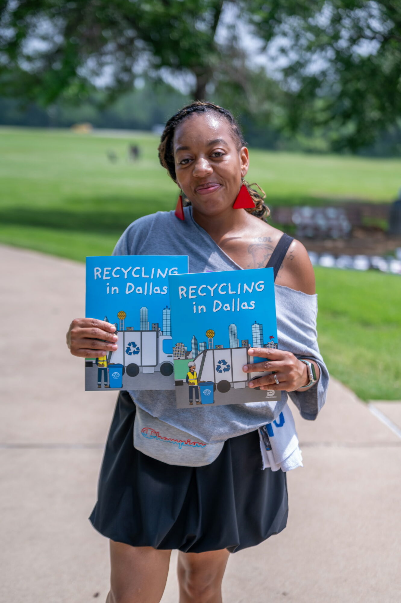 Woman holding a recycling book outdoors in a park, smiling, with trees and grass in the background.