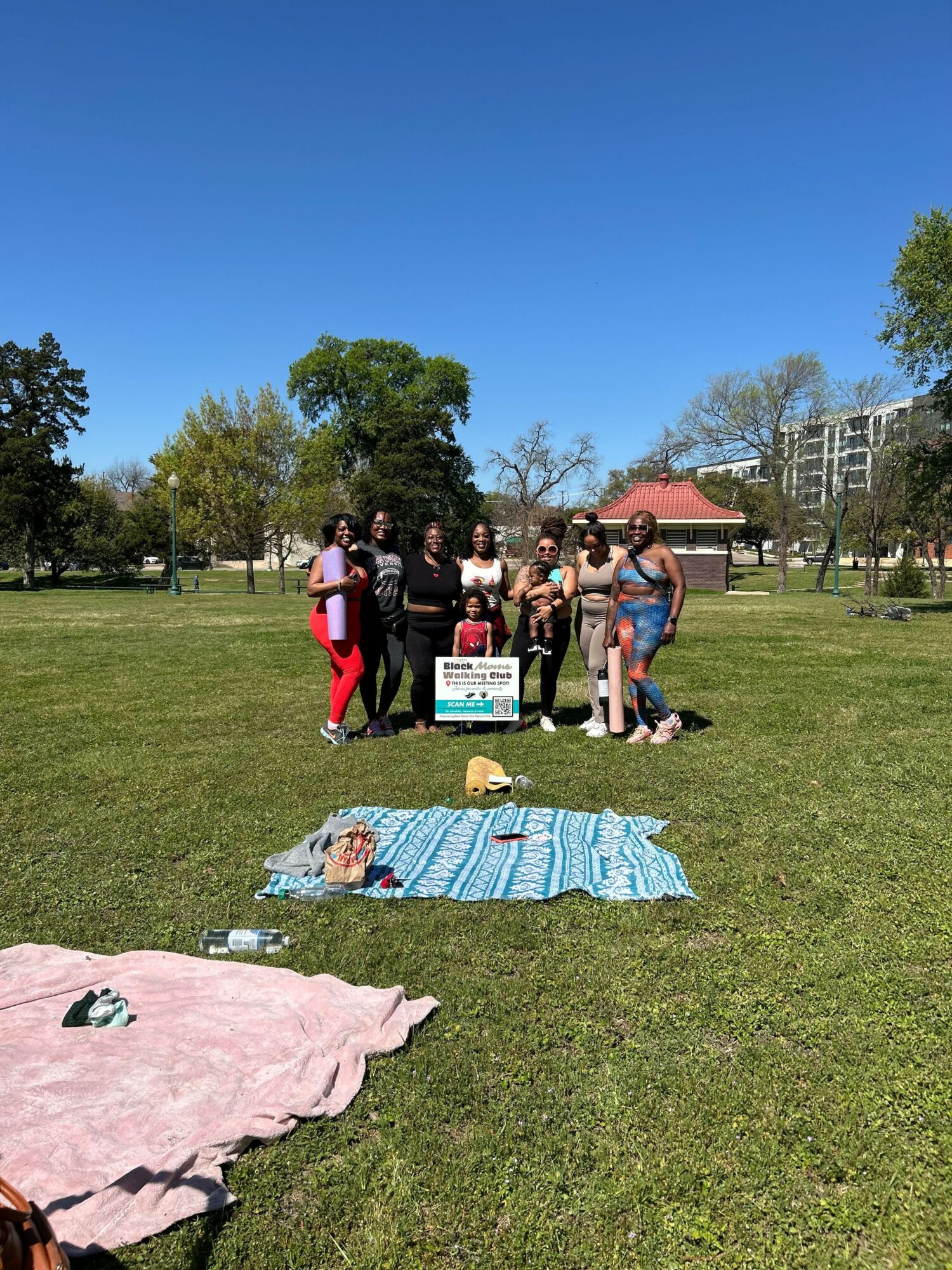 Group of people standing on grass with towels and trees in background, sunny day.