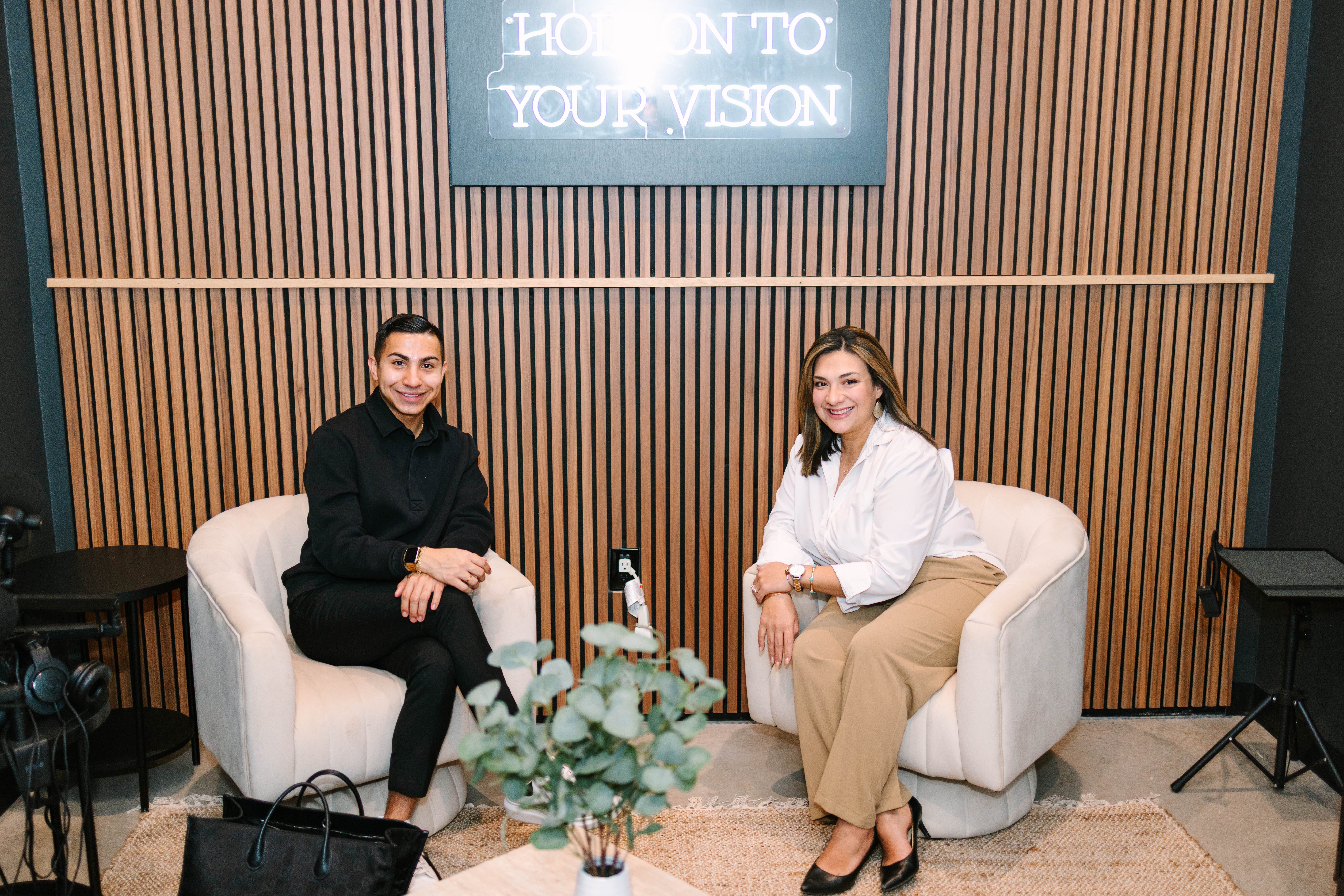 Two women sit on beige sofas in a room with wooden paneling and a sign that reads 'HOLD ON TO YOUR VISION' above them.