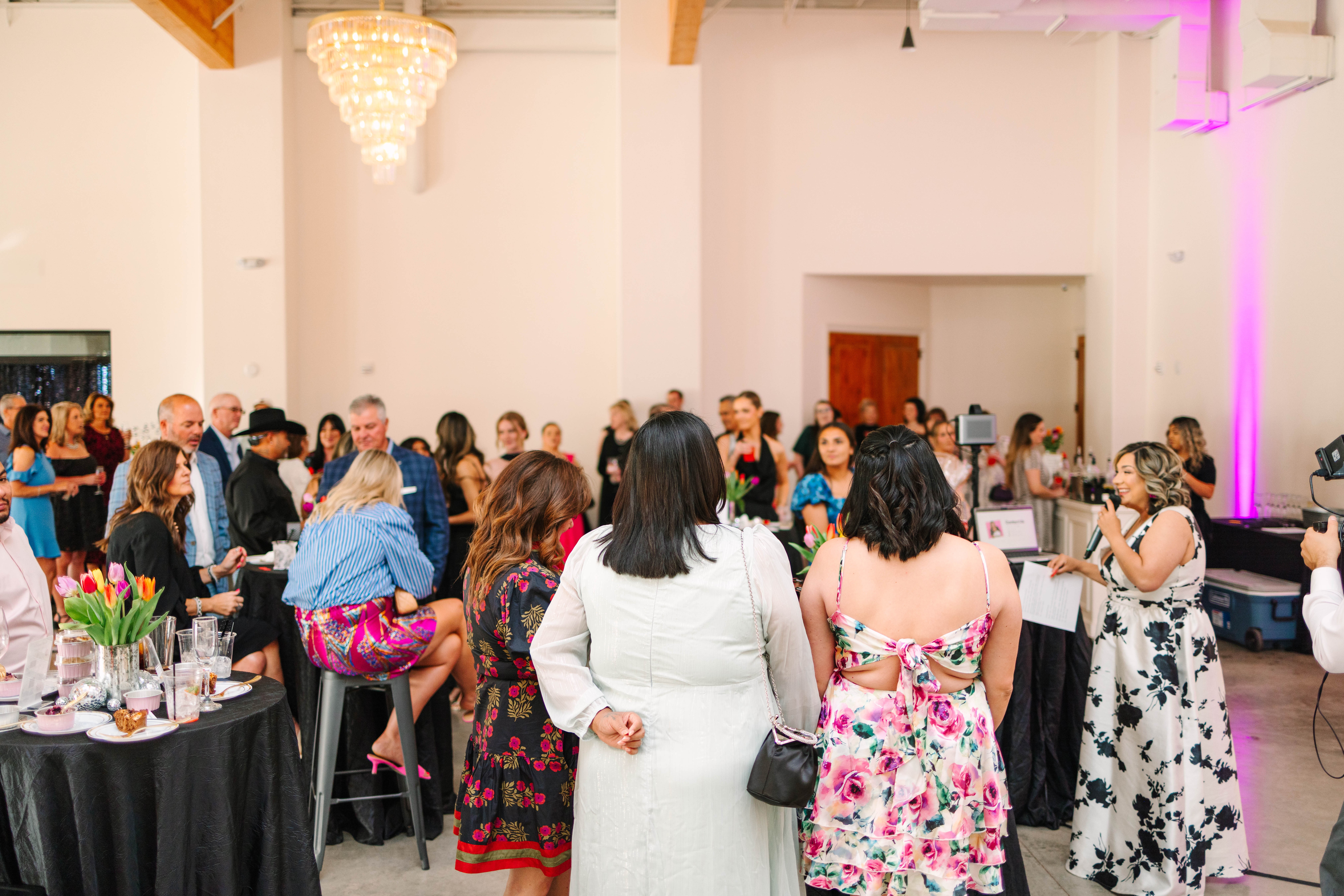 Group of people gathered in a decorated event hall, some standing and some sitting, with a table of food and flowers.