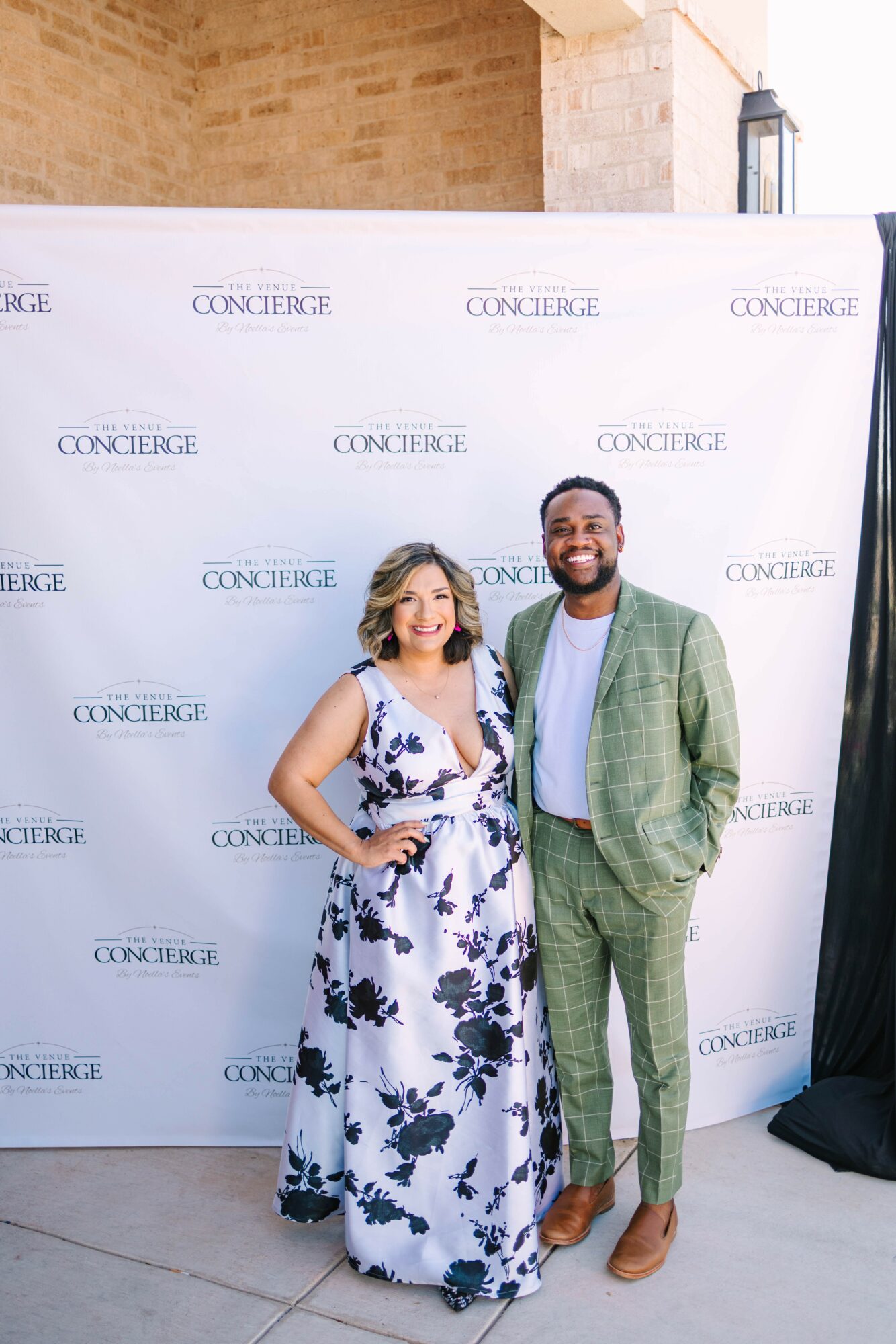 Two people standing in front of a white backdrop with 'CONCIERGE' repeated, smiling, outdoors, woman in a floral dress, man in a green suit.