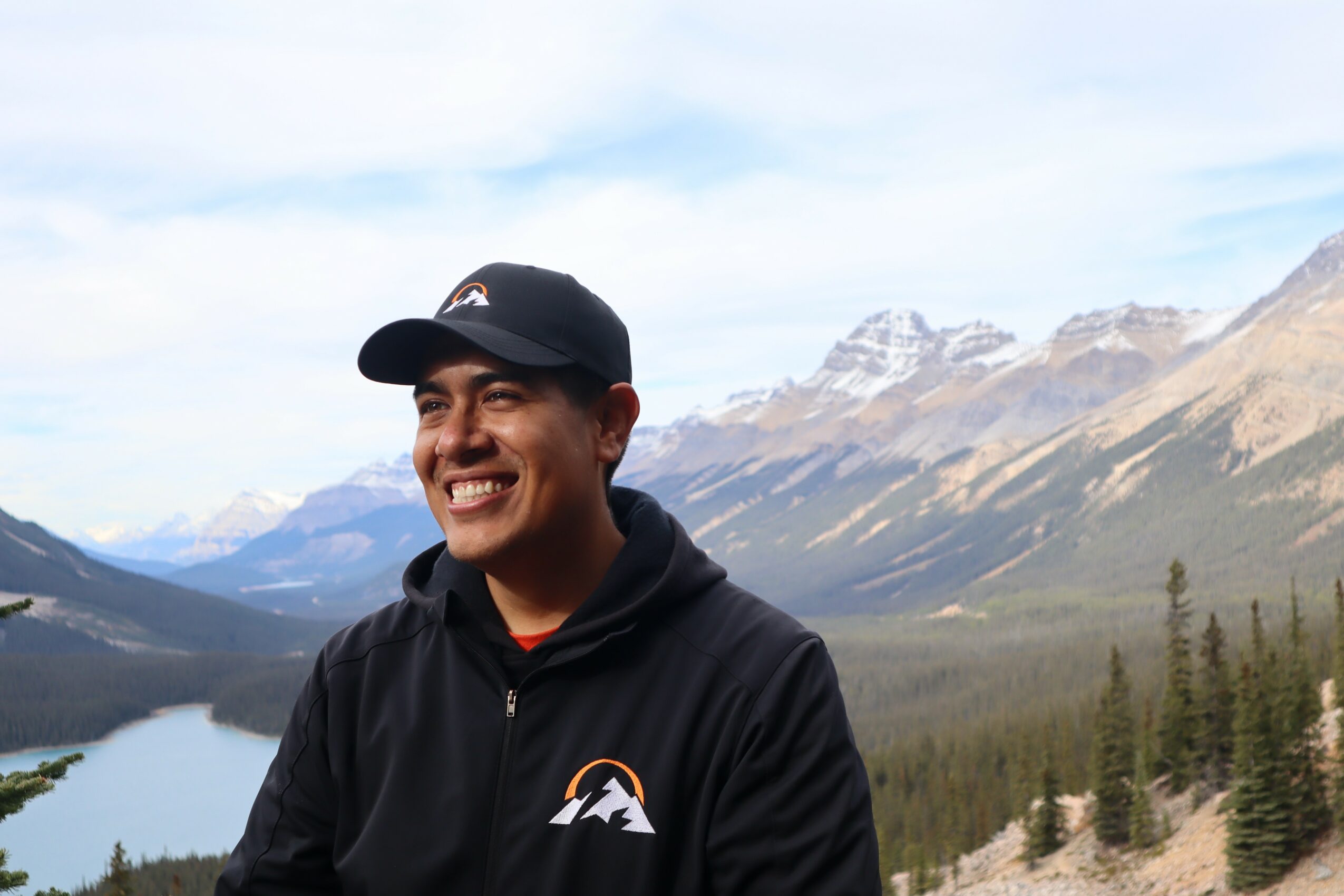 Smiling person wearing a black jacket and cap outdoors with mountains and a lake in the background.