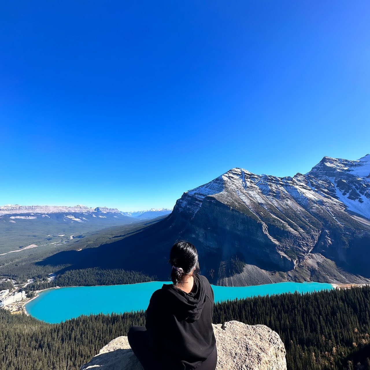 Person sitting on a rock overlooking a mountain lake with snow-capped peaks and a clear blue sky.