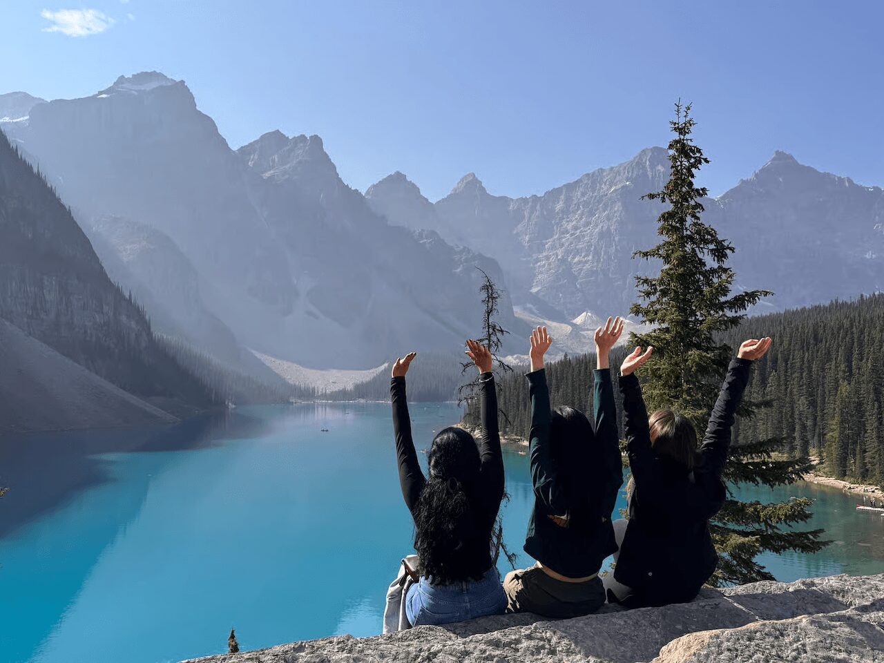 Three people sit on rocks by a lake with mountains and trees in the background, raising their arms.