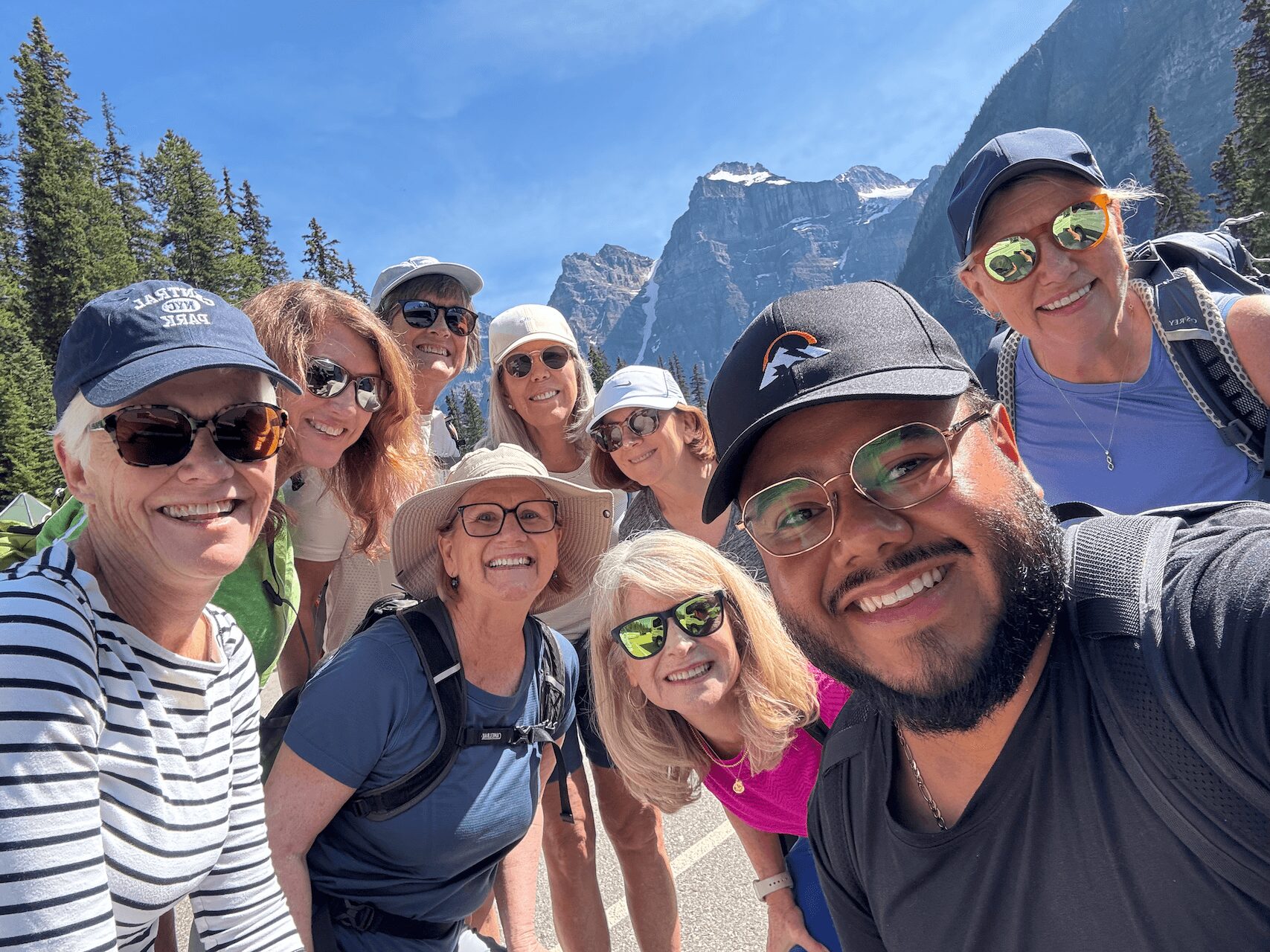 Group of nine people outdoors in a mountainous area, smiling, wearing hats and sunglasses, with trees and mountains in the background.