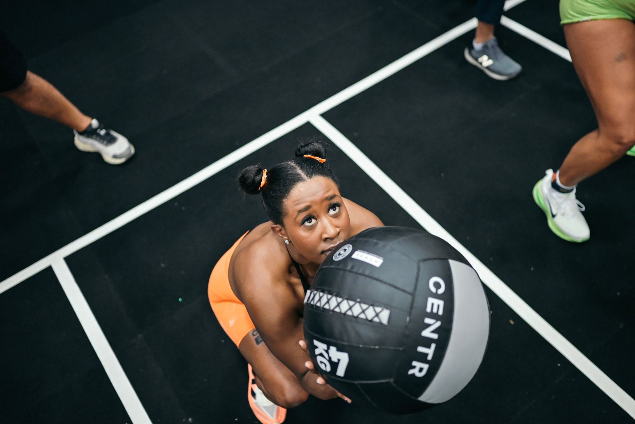 Woman holding a medicine ball on a gym floor, surrounded by other people exercising.
