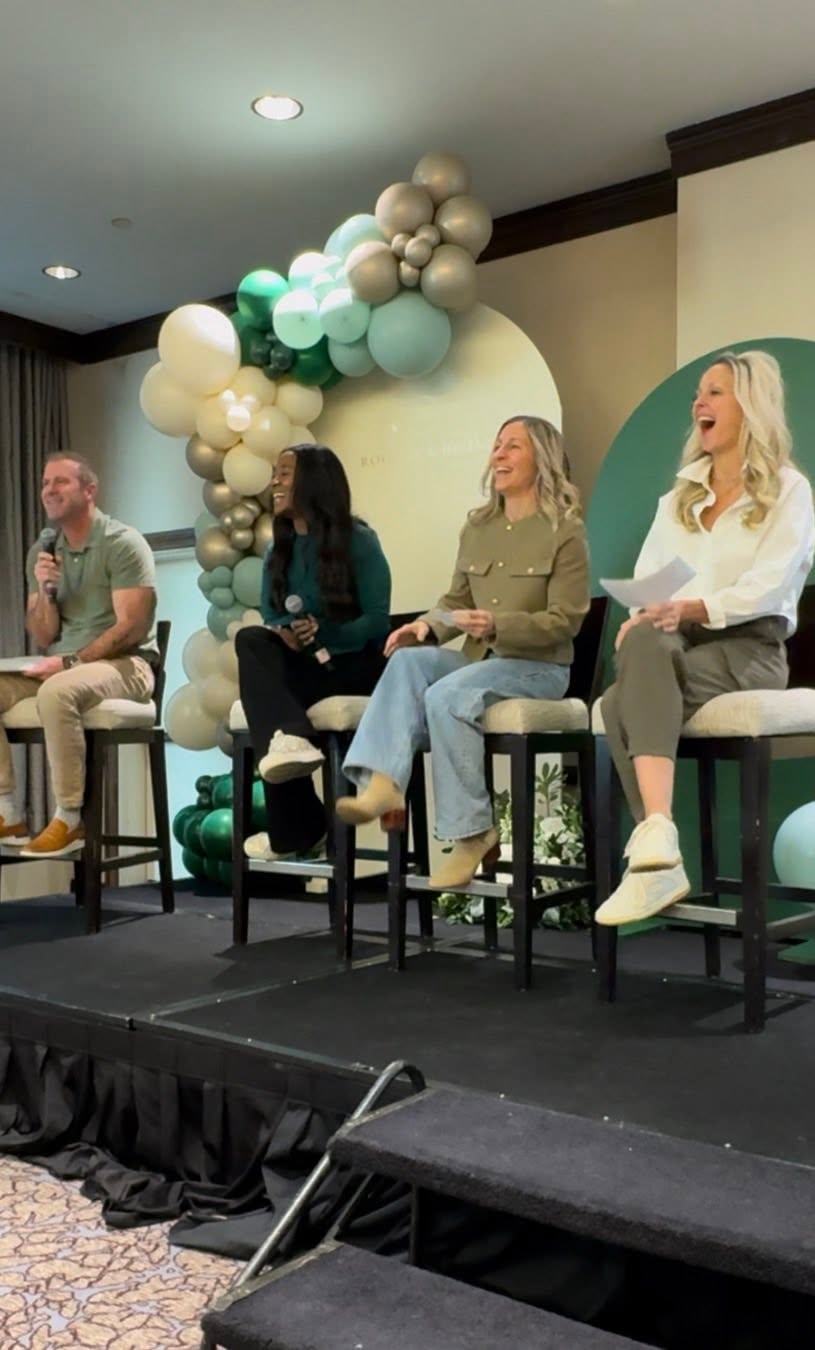 Four people sit on stage with a balloon arch behind them, engaged in a panel discussion or presentation.