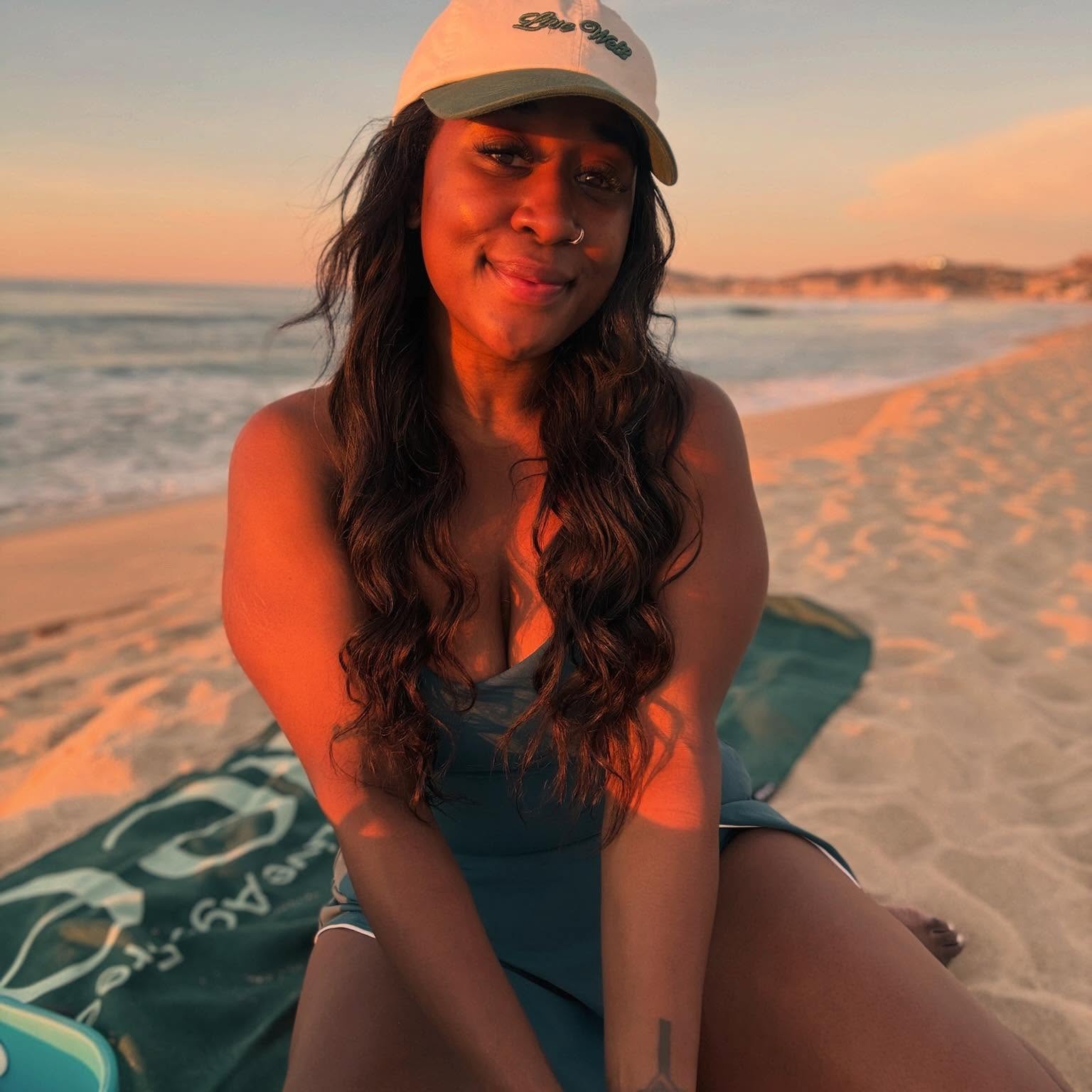 Woman sitting on a beach at sunset, smiling, wearing a white cap and a dark top.
