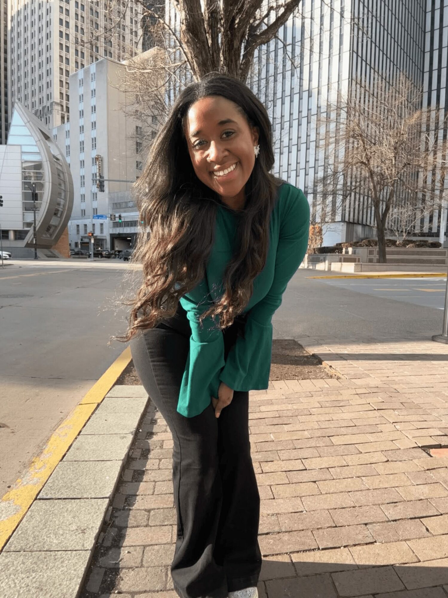 Young woman with long dark hair smiling outdoors in cityscape, wearing green top and black pants.