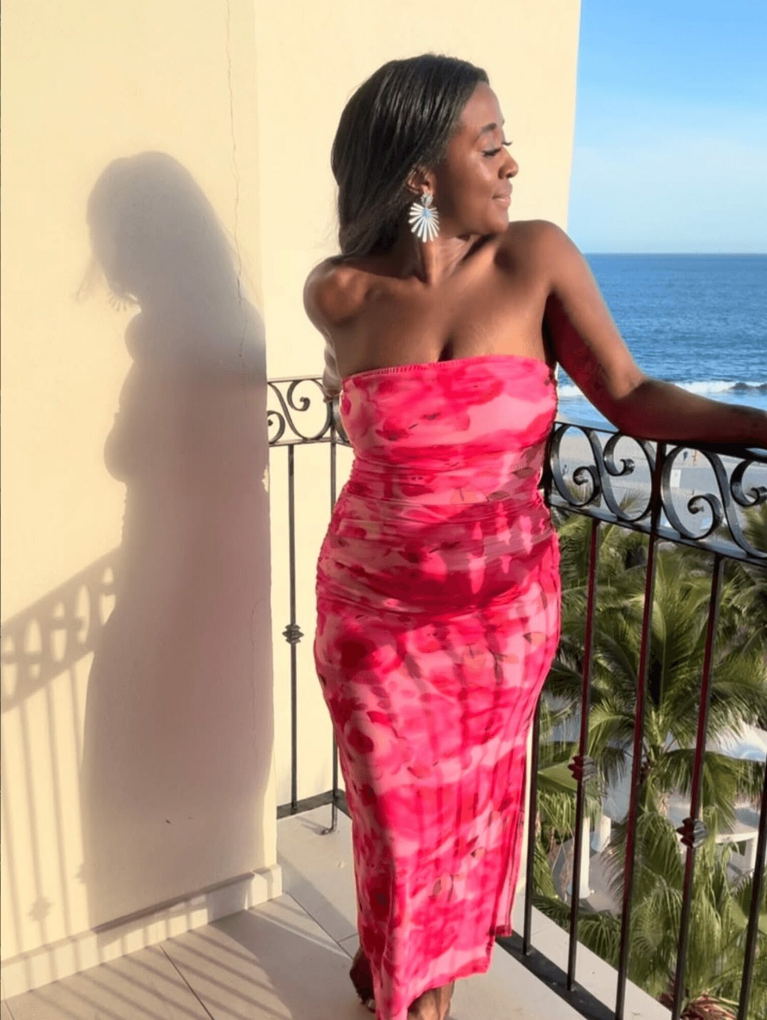 Woman in a pink strapless dress standing on a balcony with ocean view, sunlight casting her shadow on the wall.