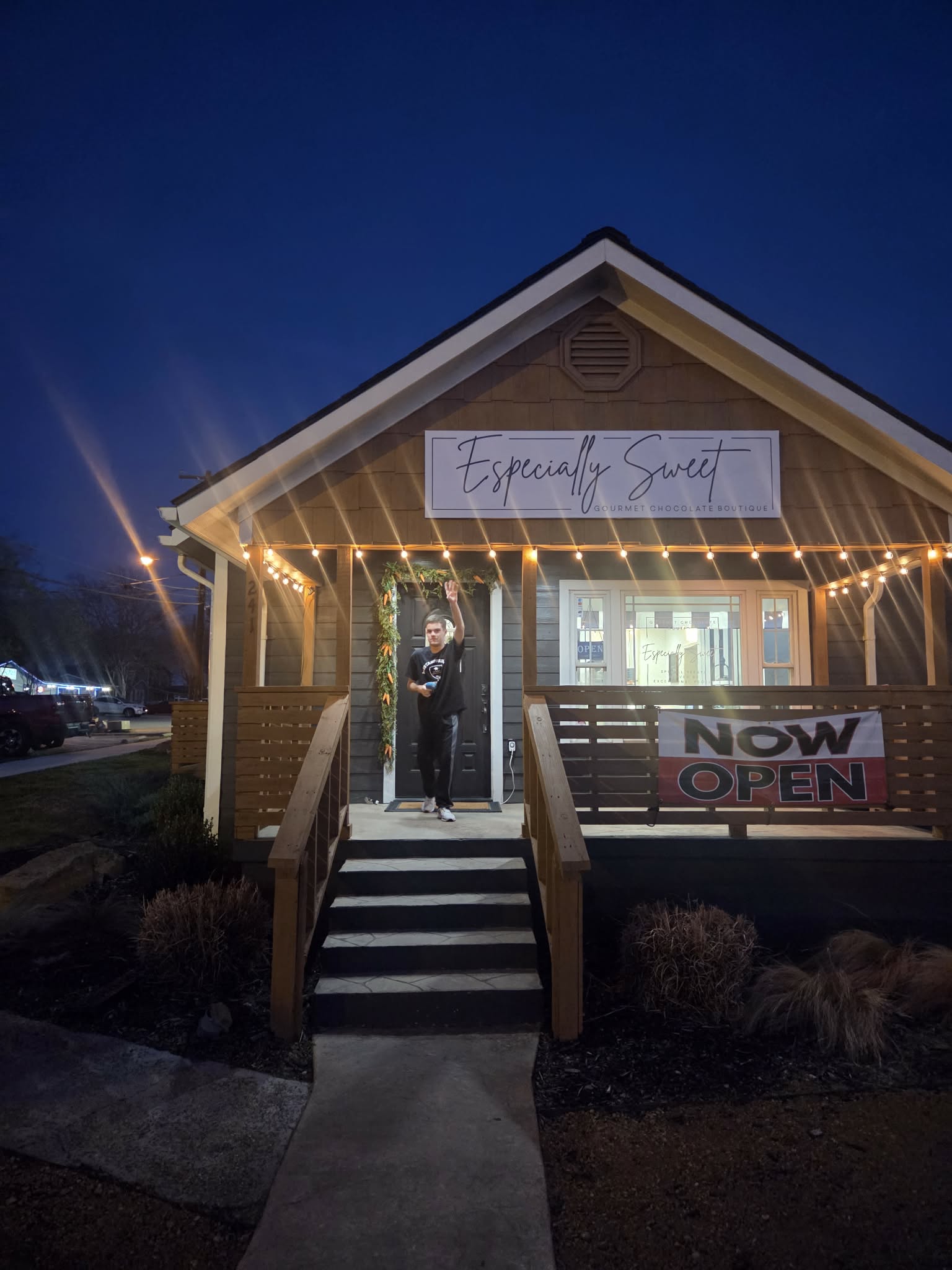 Small building with string lights, a person standing at the entrance, and signs indicating it is now open.