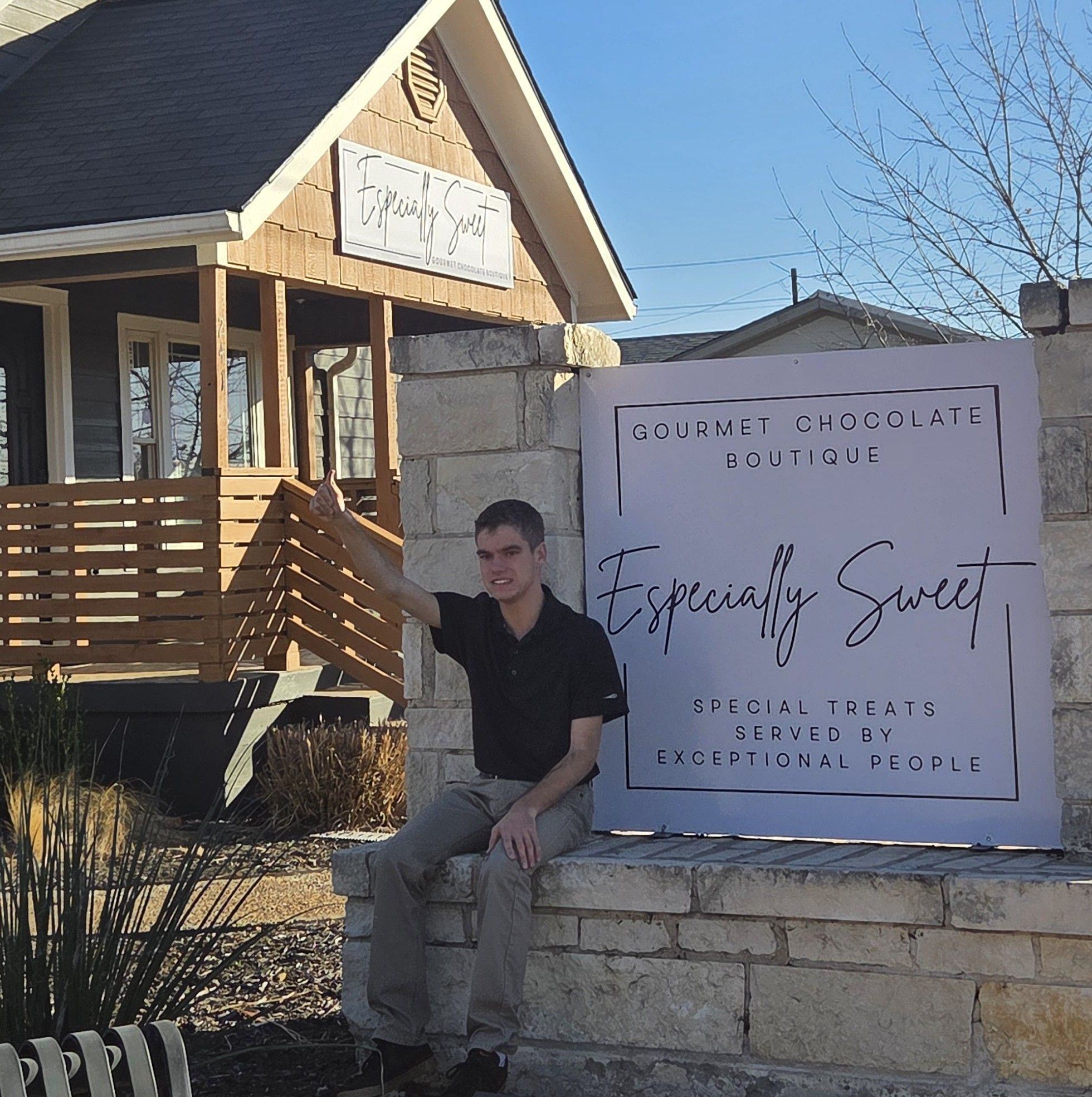 Young person sitting on a stone ledge next to a large sign outside a boutique, with a house in the background.