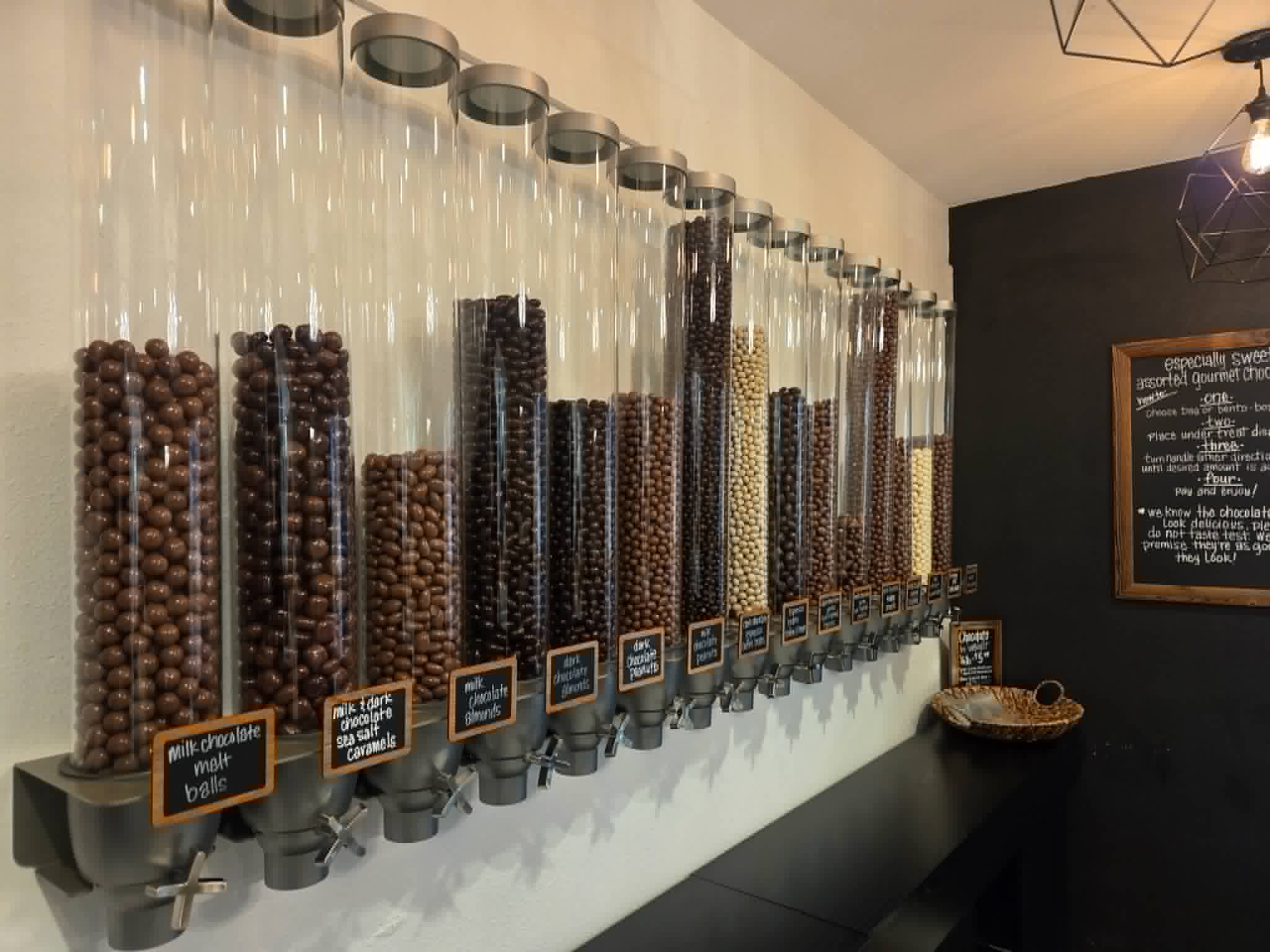 Wall with multiple tall glass containers filled with different types of coffee beans, each labeled with small signs, in a cafe setting.