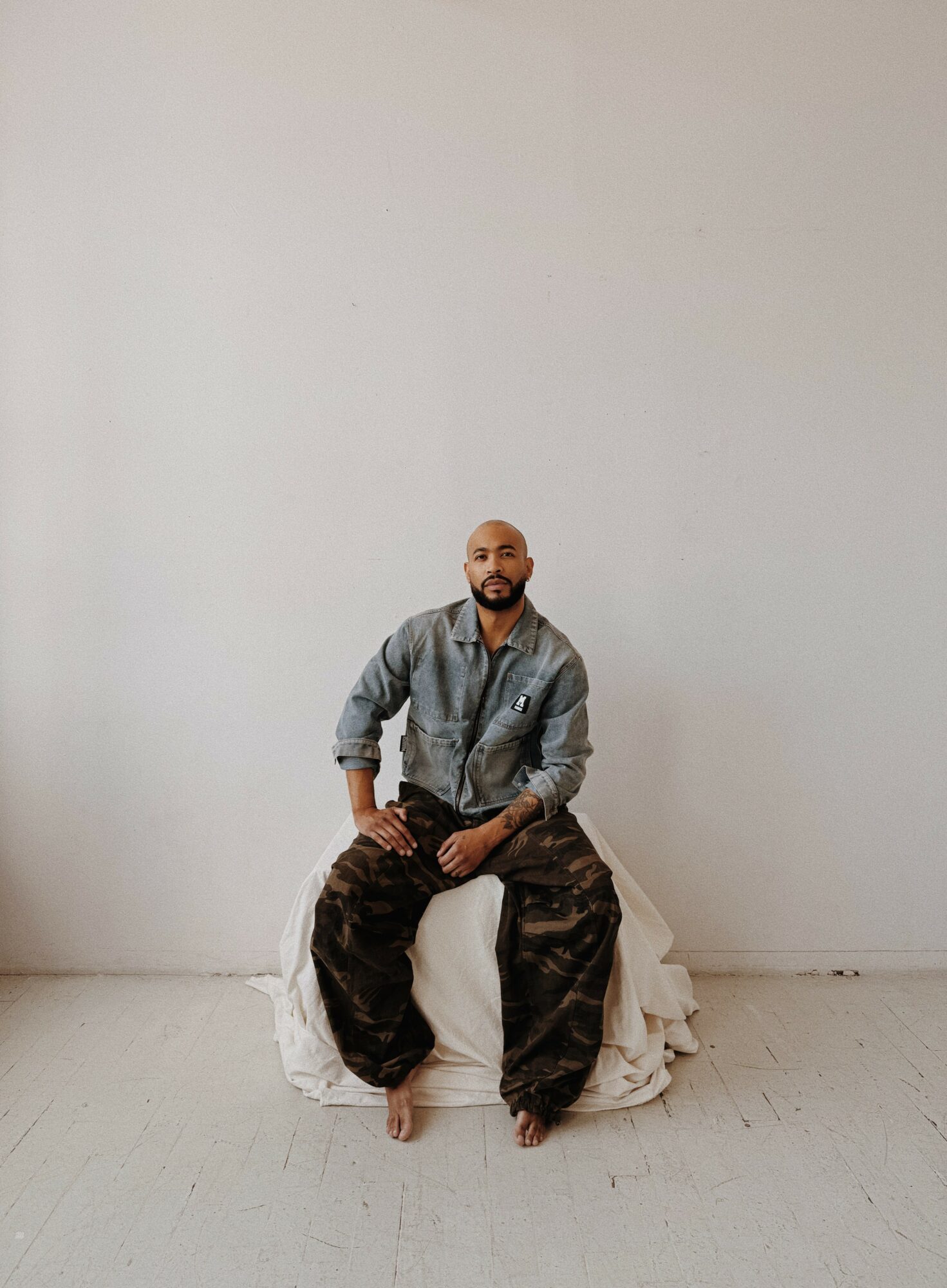 Man sitting on a white cloth against a plain white wall, wearing a gray jacket and camouflage pants.