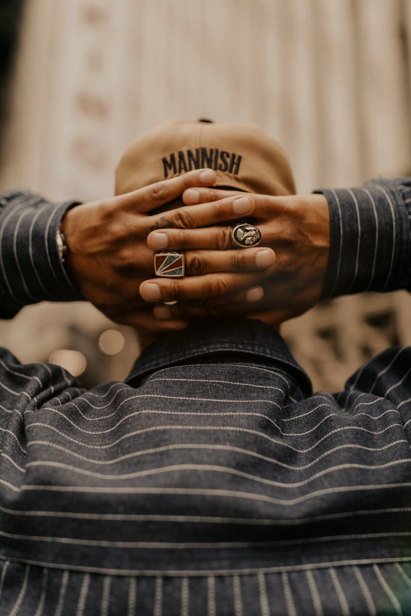 Person wearing a striped shirt holds a tan cap with 'MANNISH' written on it, hands with rings visible, background blurred.
