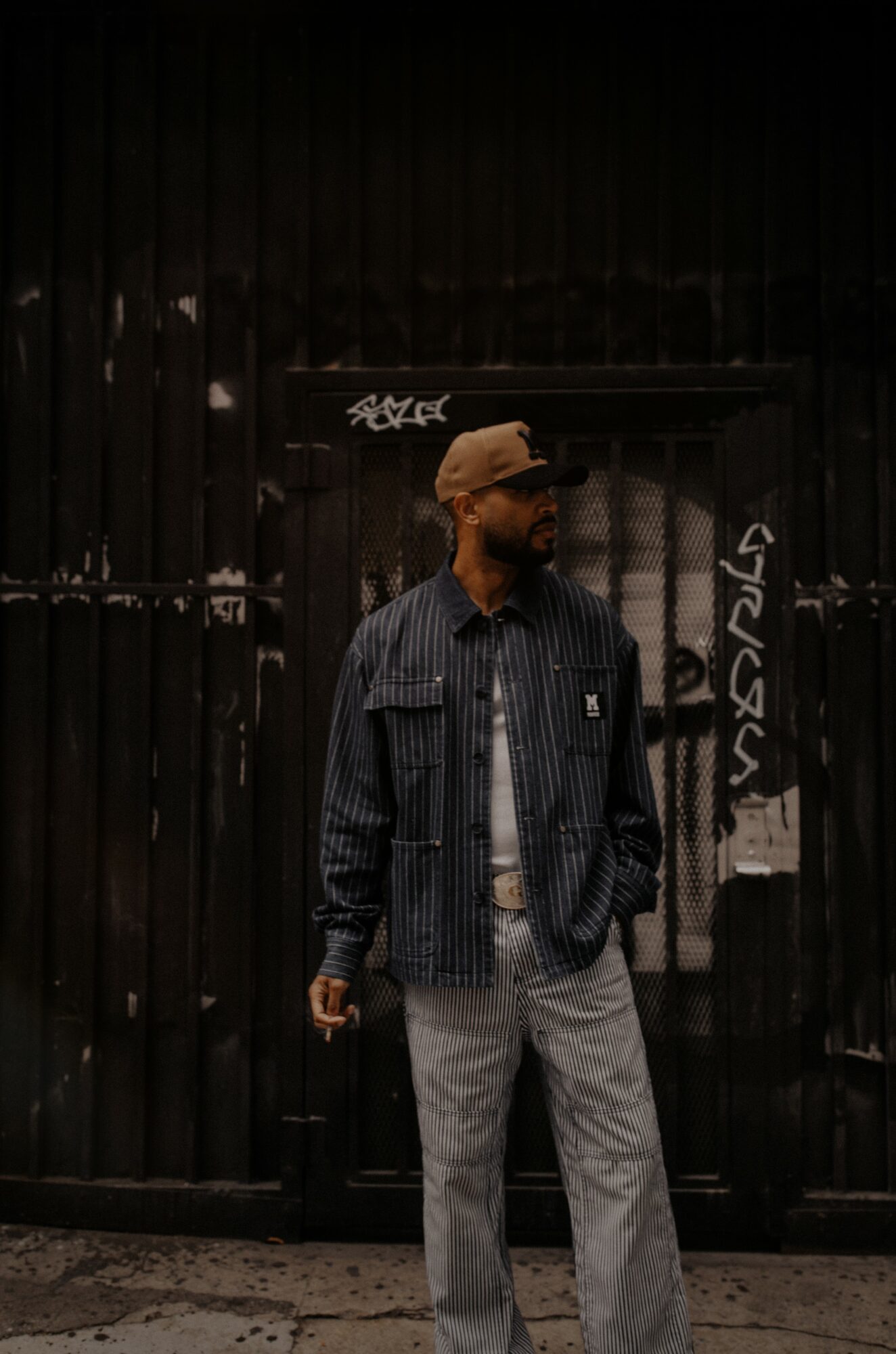 Man standing in front of a black gate, wearing a jacket, striped pants, and a cap, holding a cigarette.