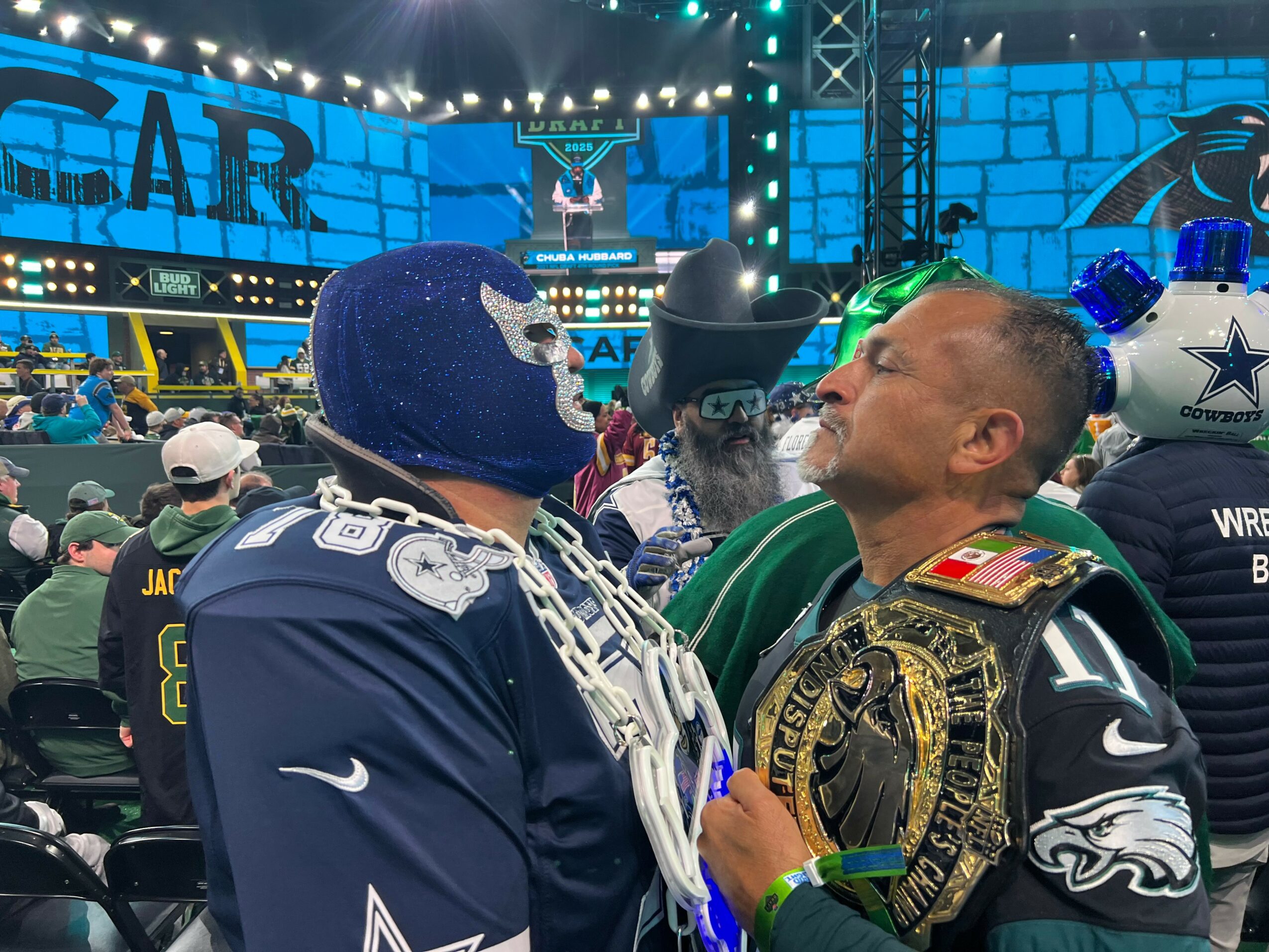 Two men, one in a blue sports jersey and mask, the other in a black sports uniform with a championship belt, face each other at a stadium.