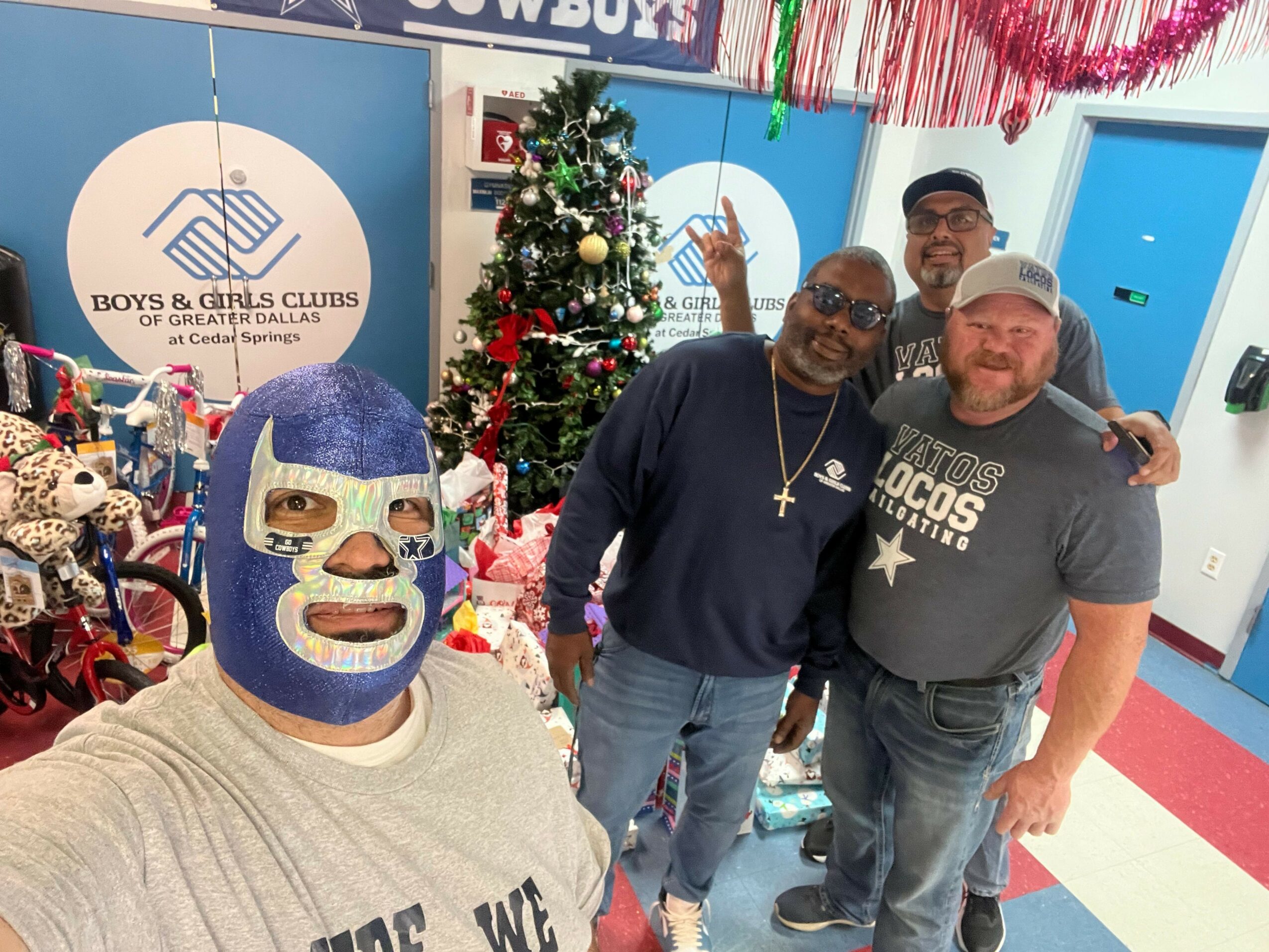 Four people posing in front of a Christmas tree and a sign, with one person wearing a blue mask and glasses.