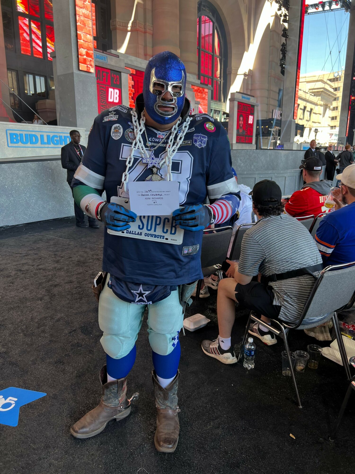 Person dressed as a superhero with a blue mask, cape, and chain, walking on a city street with people and buildings in background.