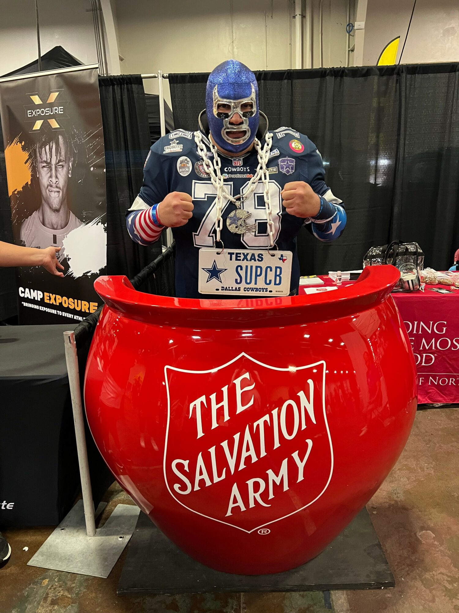 Person dressed as a clown with face paint and a blue hat, standing behind a red donation barrel labeled 'The Salvation Army'.