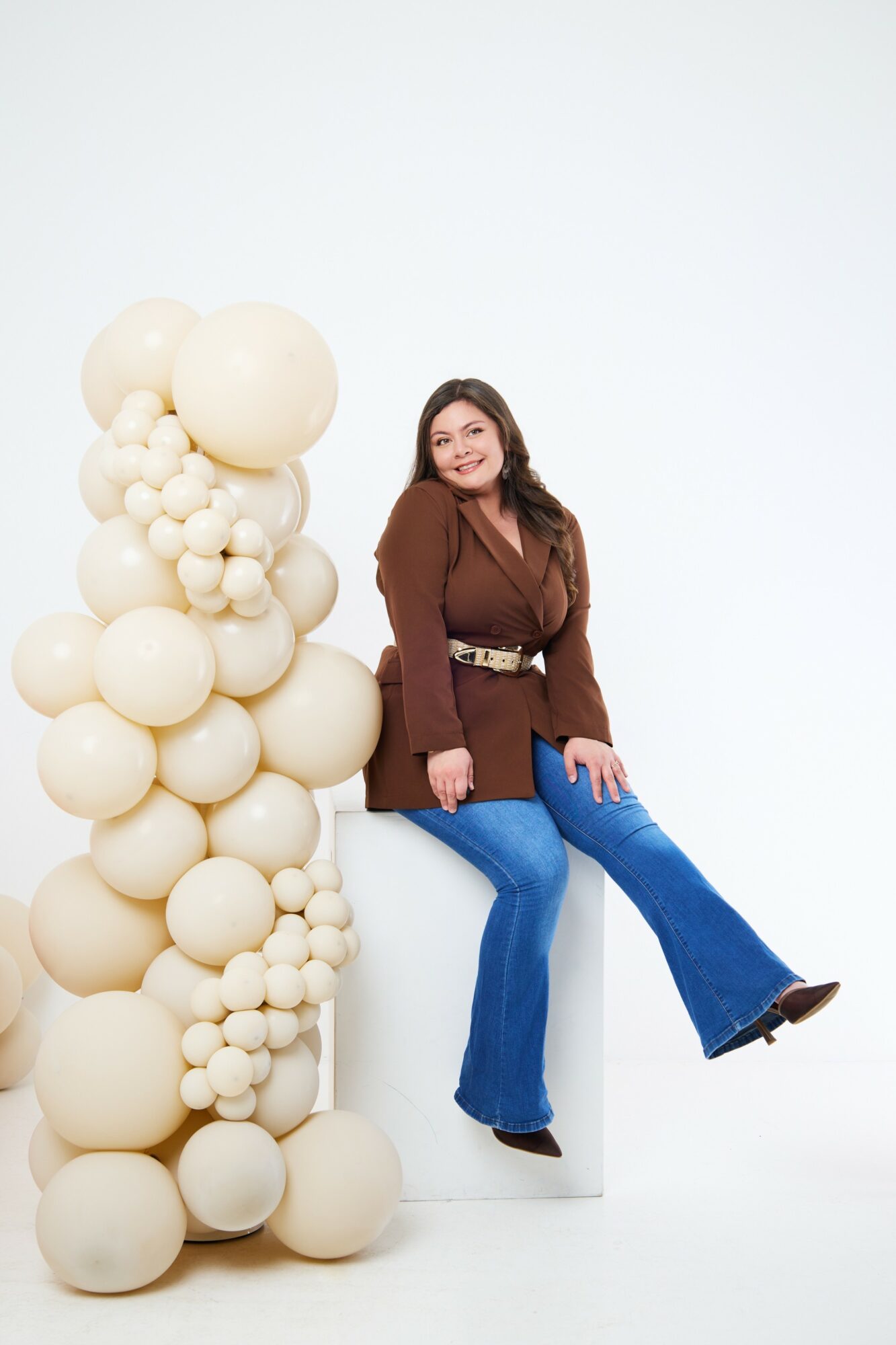 Woman with long hair, brown top, and blue jeans sitting on a white block, smiling, next to a large cluster of cream-colored balloons.