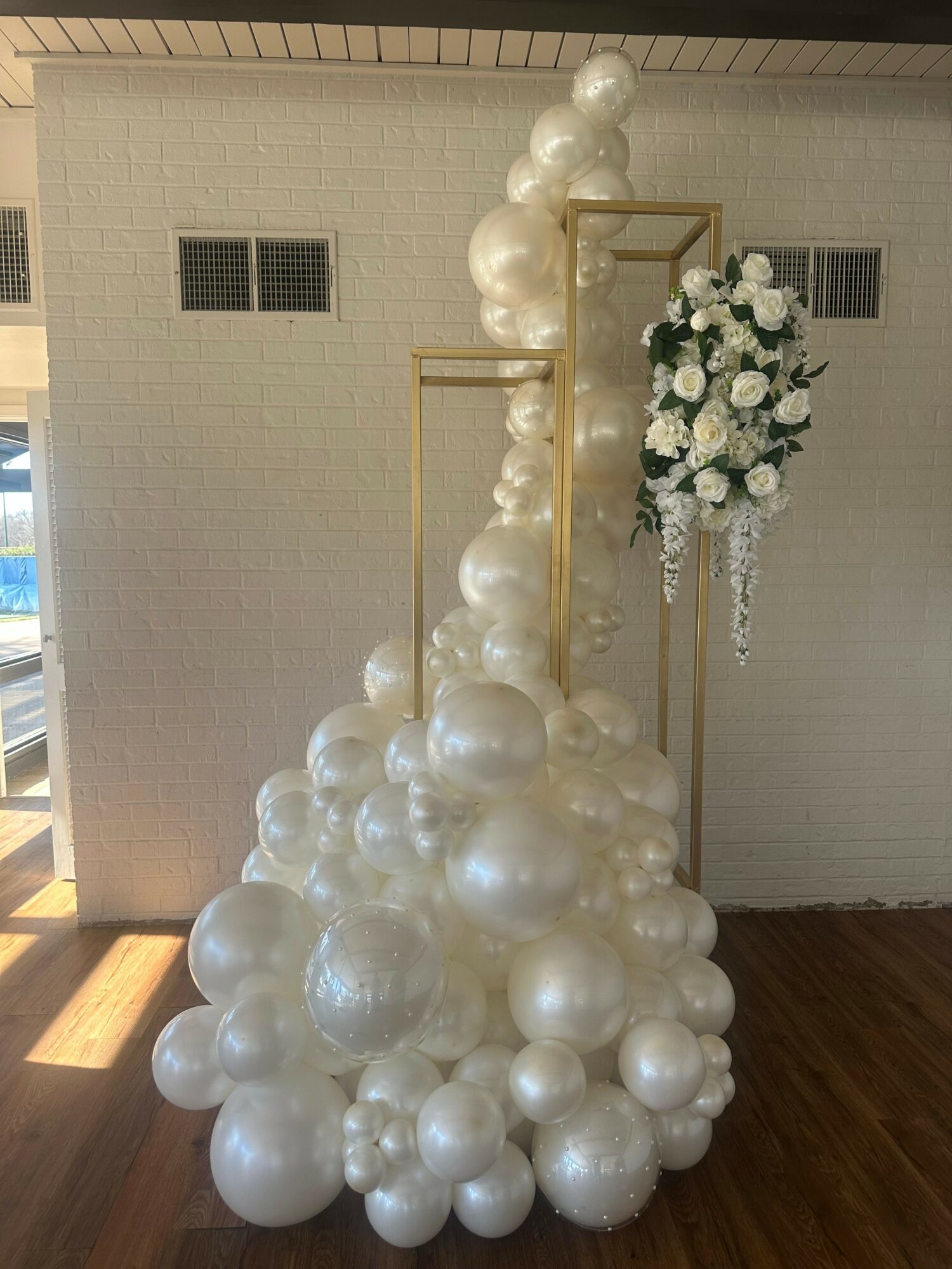 Decorative arrangement with white balloons and a floral centerpiece on a gold stand, set against a plain wall.