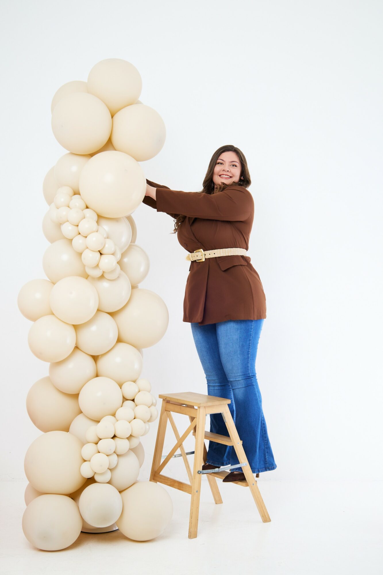 Woman standing on a stool, reaching towards a tall arrangement of white balloons, smiling, against a plain white background.
