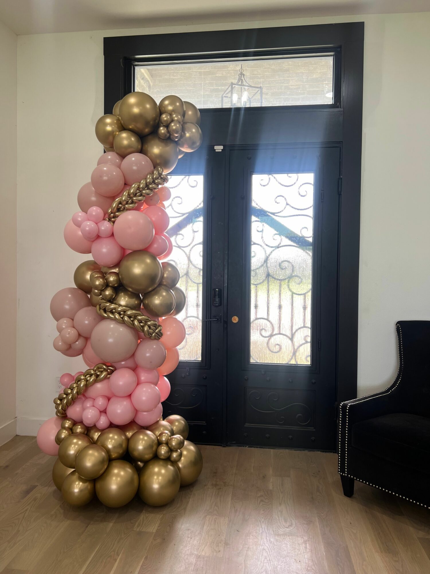 Decorative balloon arrangement with pink and gold balloons, some with metallic finish, in front of a black door with glass panels.