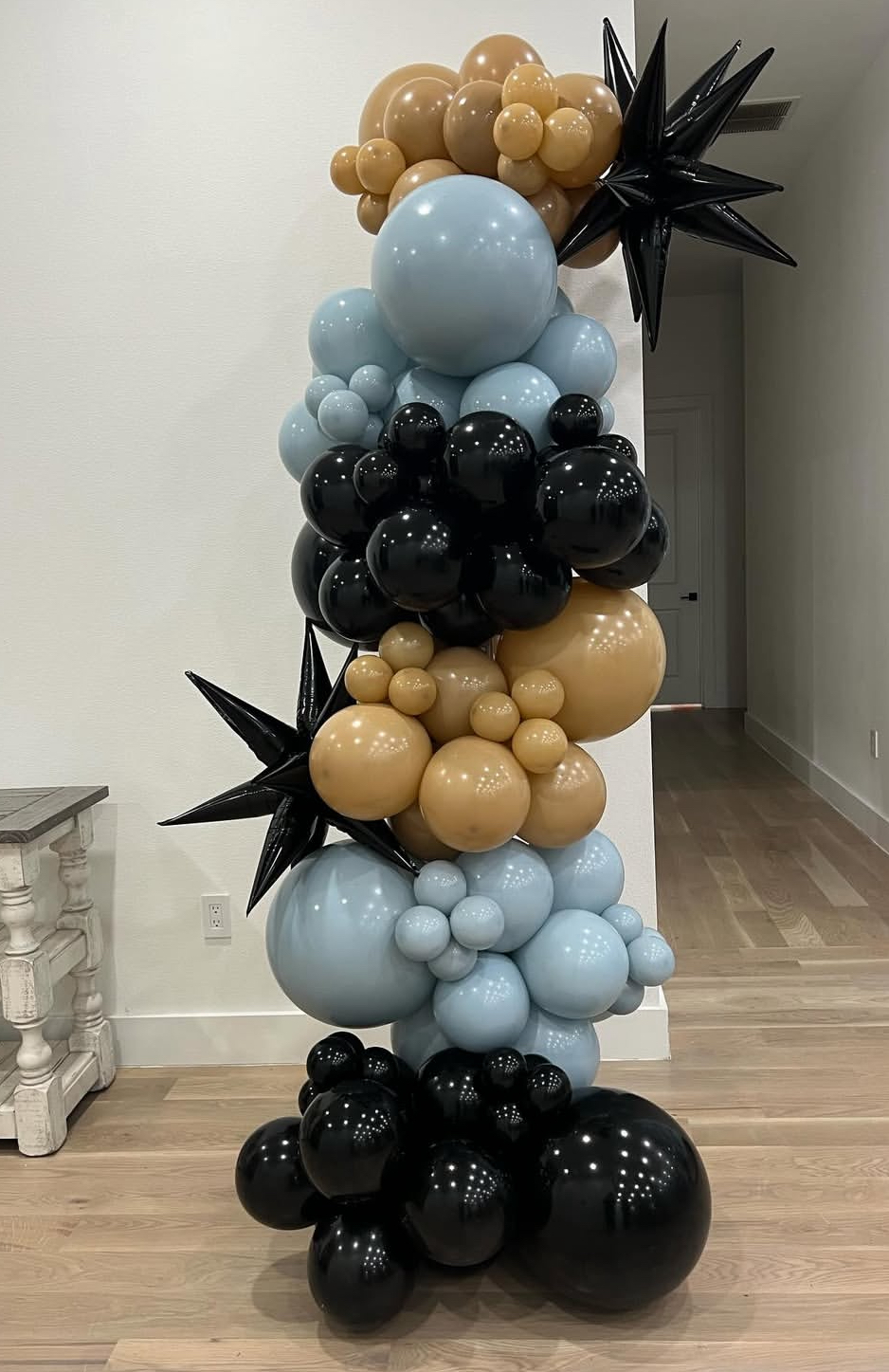 Vertical balloon arrangement with beige, black, and light blue balloons, including star-shaped balloons, in a room with wooden floor.