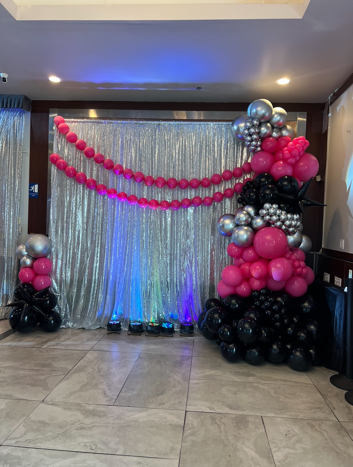 Decorative balloon arrangement with pink, black, silver, and gray balloons in front of a shiny silver curtain backdrop, with pink balloon garlands overhead.