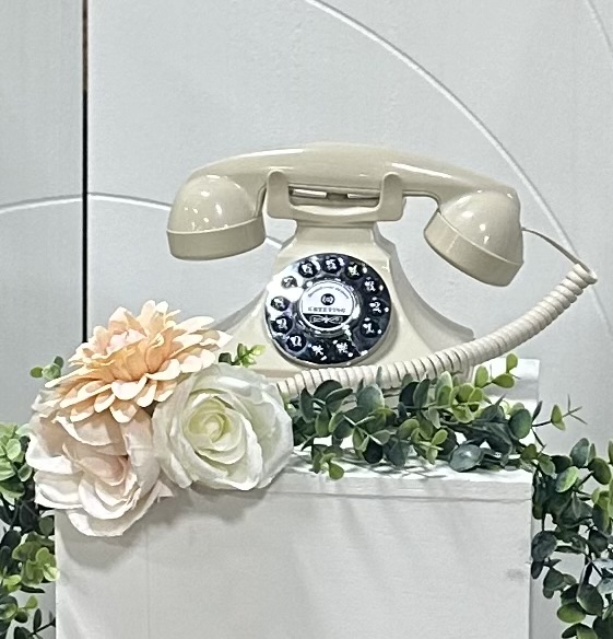 Vintage rotary telephone with flowers and greenery on a white tiled wall, no rotation needed.