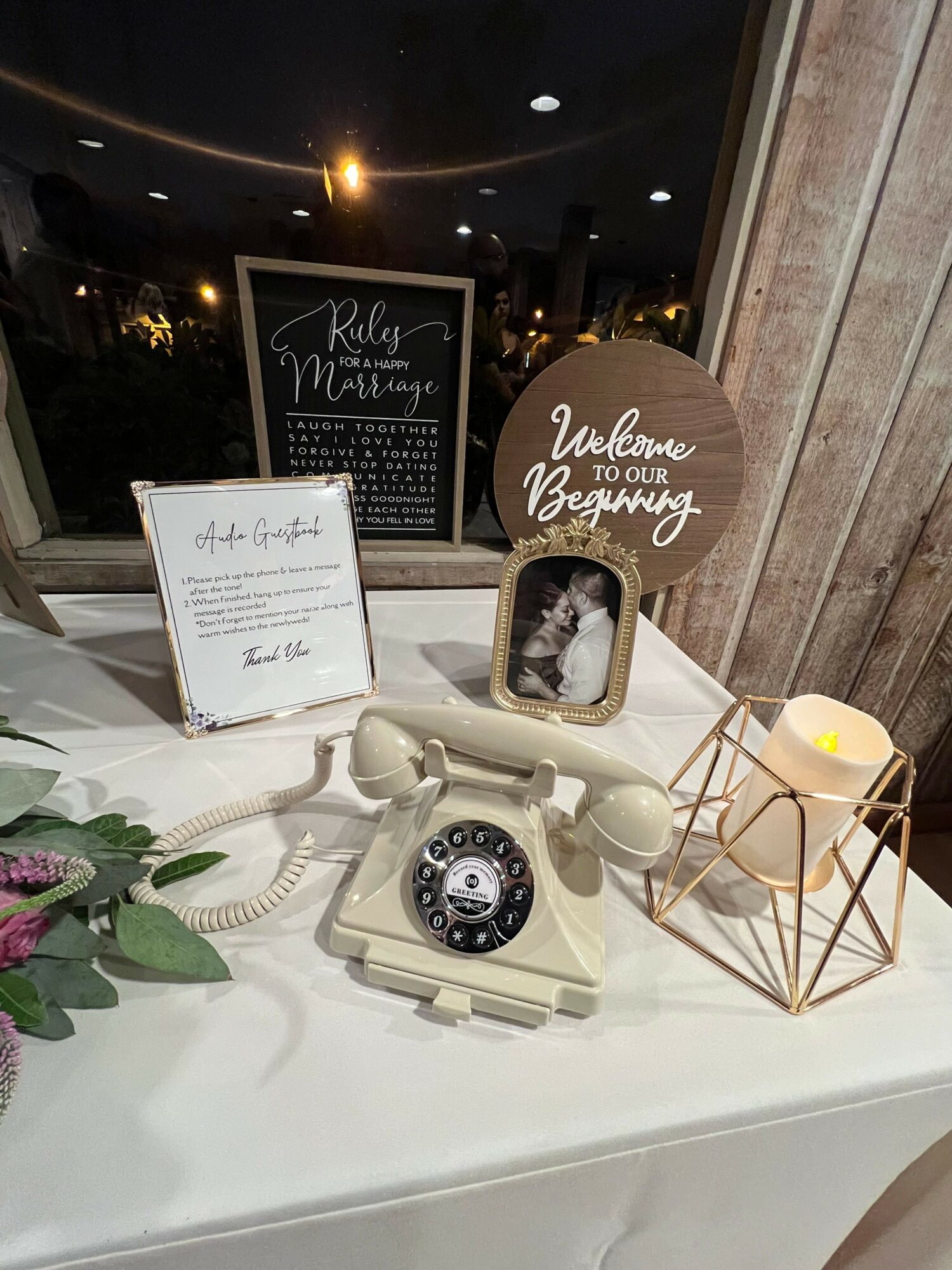 Decorative table with vintage rotary phone, framed photo, chalkboard sign, and geometric candle holder, against a wooden wall background.