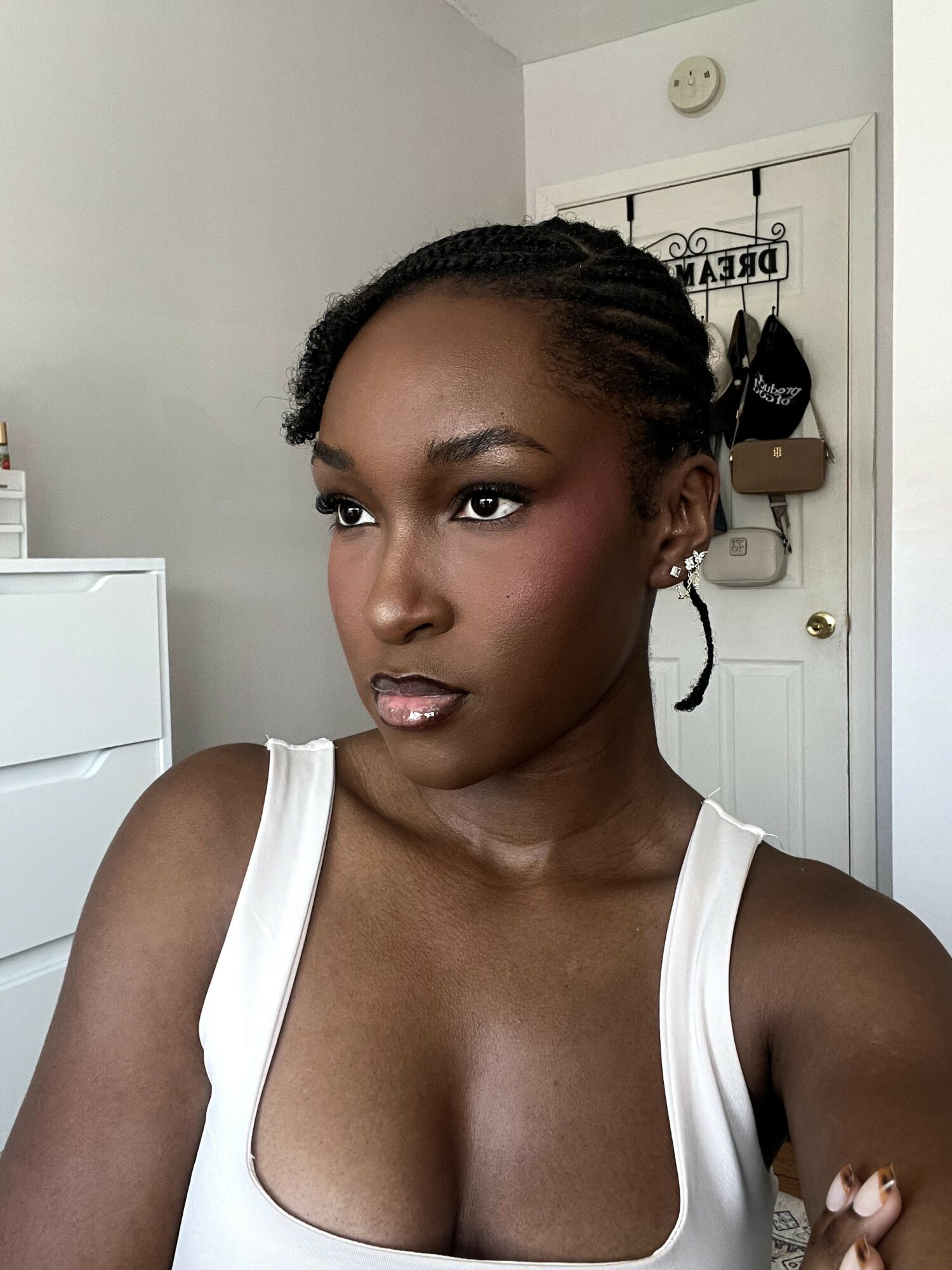 Portrait of a woman with braided hair wearing a white tank top, looking to the side in a room with a door and wall decor
